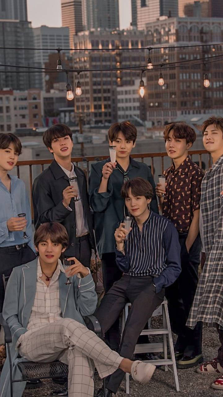 Bts Aesthetic 桌布 壁紙 — 七位穿著時尚的年輕男子在屋頂上放鬆，背景是城市天際線，在黃金時刻燈串閃爍。