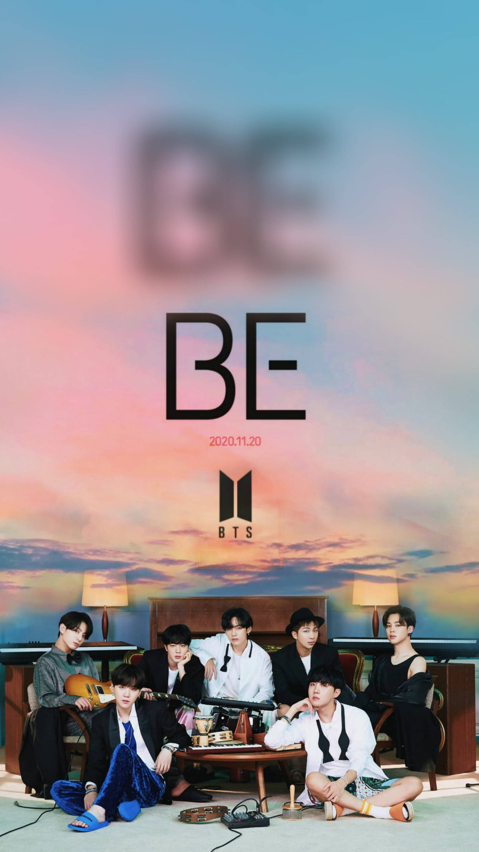 BTS 桌布 壁紙 — 七位穿著時尚的年輕男子在溫暖照明的房間內休閒地坐著，背景是充滿活力的日落天空和巨大的「BE」文字。