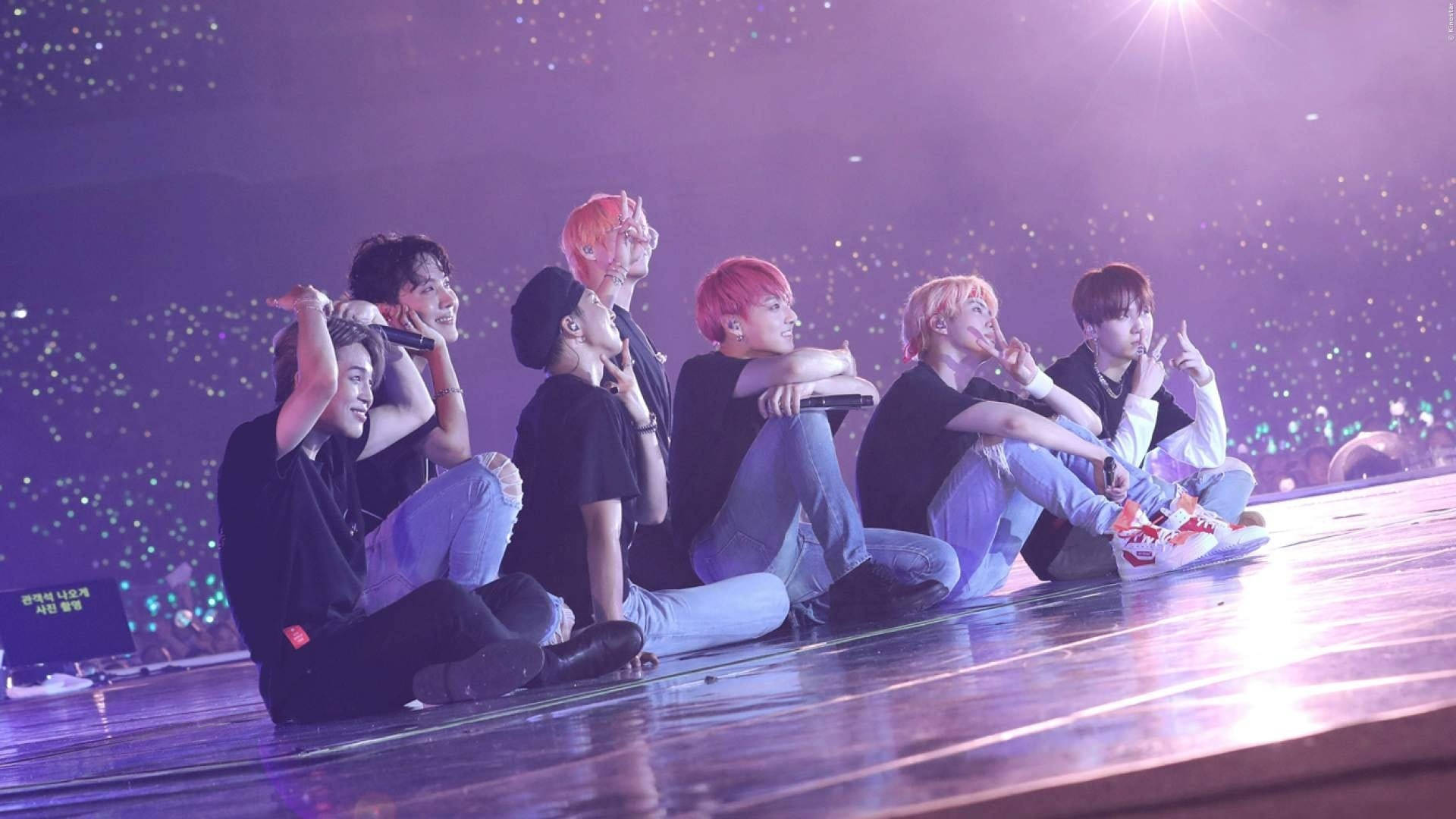 BTS 桌布 壁紙 — 七位表演者在紫色燈光下，隨意地坐在一個閃亮的舞台上，背景是閃爍的觀眾。