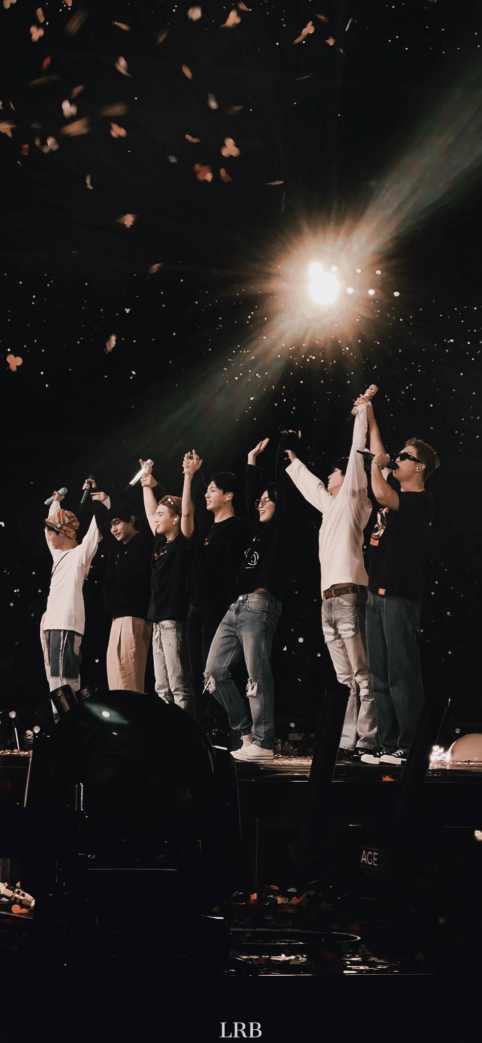 bts iPhone 桌布 壁紙 — 七位表演者在舞台上於明亮聚光燈下舉起手臂，紙屑在黑暗的音樂會場景中飄落。