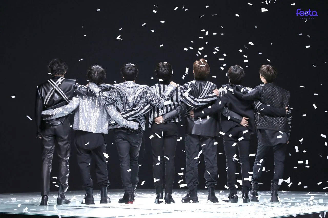 Bts Concert 桌布 壁紙 — 七位表演者在舞台上背對背站立，手臂相連，白色紙屑在明亮燈光下落在他們周圍。
