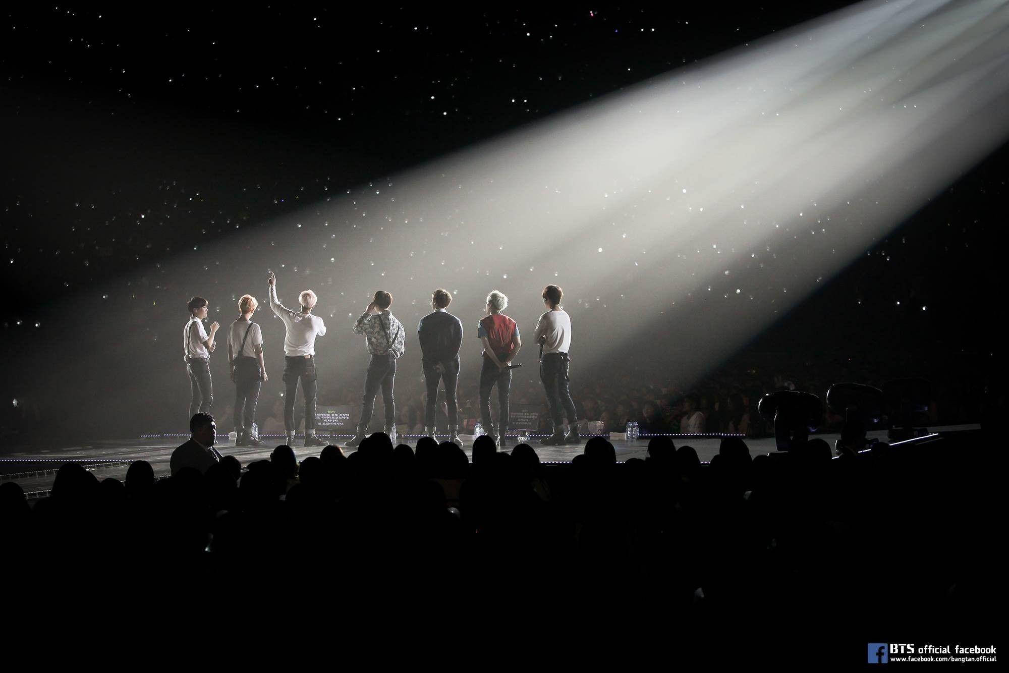 BTS 黑色 桌布 壁紙 — 七位表演者站在漆黑的舞台上，沐浴在單一明亮的聚光燈下，背景有星星，觀眾以剪影形式觀看。