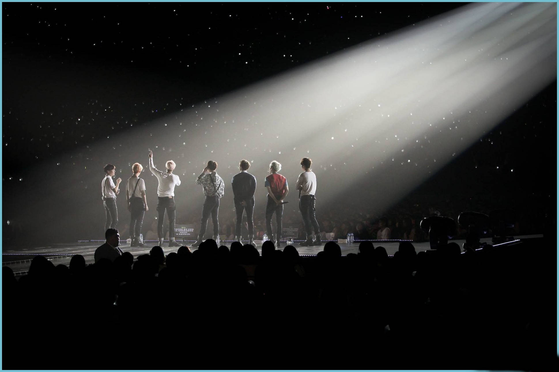 BTS 桌布 壁紙 — 七位表演者站在燈光昏暗的舞台上，沐浴在單一聚光燈下，背景是星空，下方觀眾形成剪影。