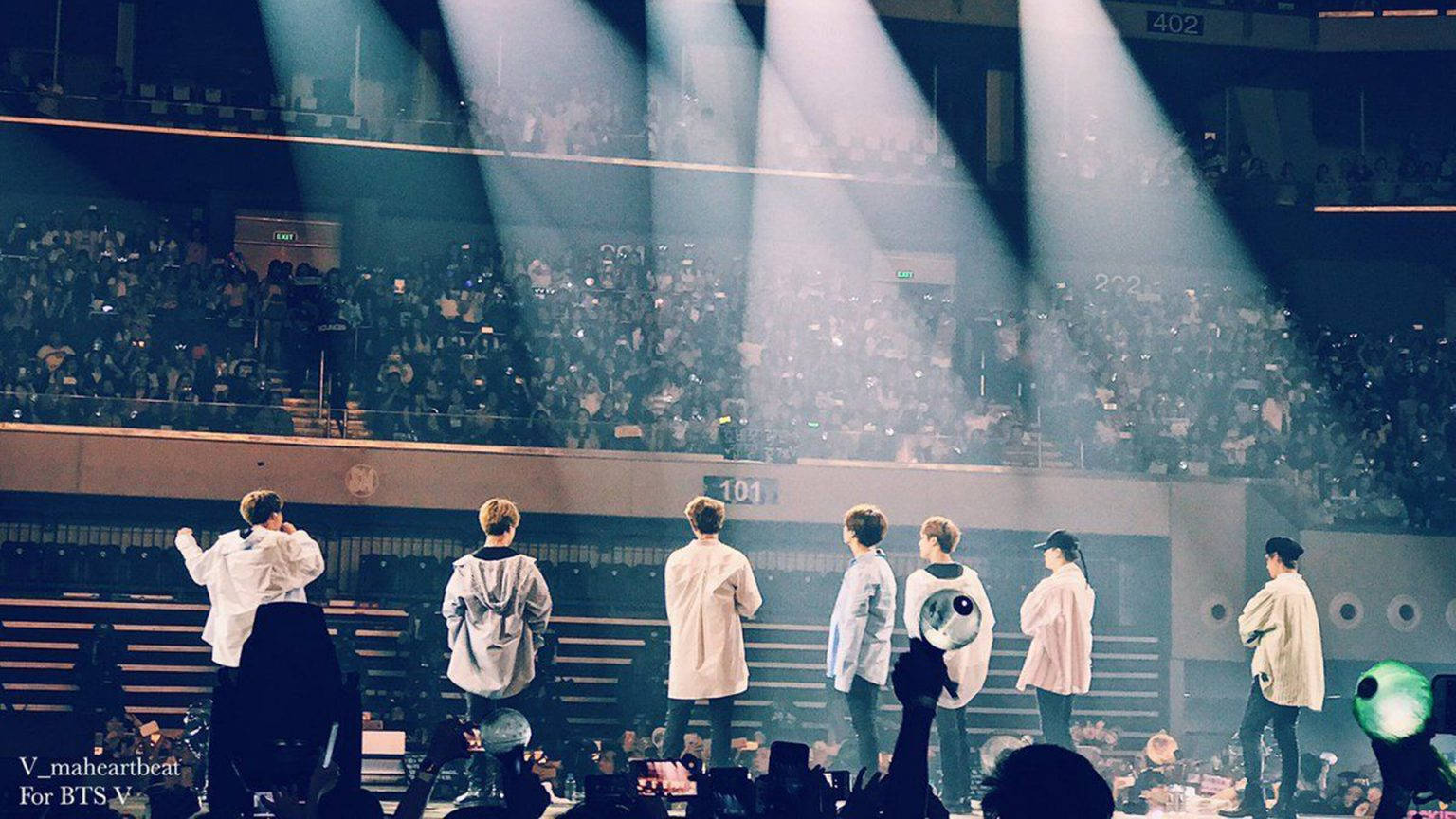 BTS 桌布 壁紙 — 七位表演者站在燈光璀璨的舞台上，面對擠滿場館的龐大觀眾，在戲劇性的聚光燈下演出。