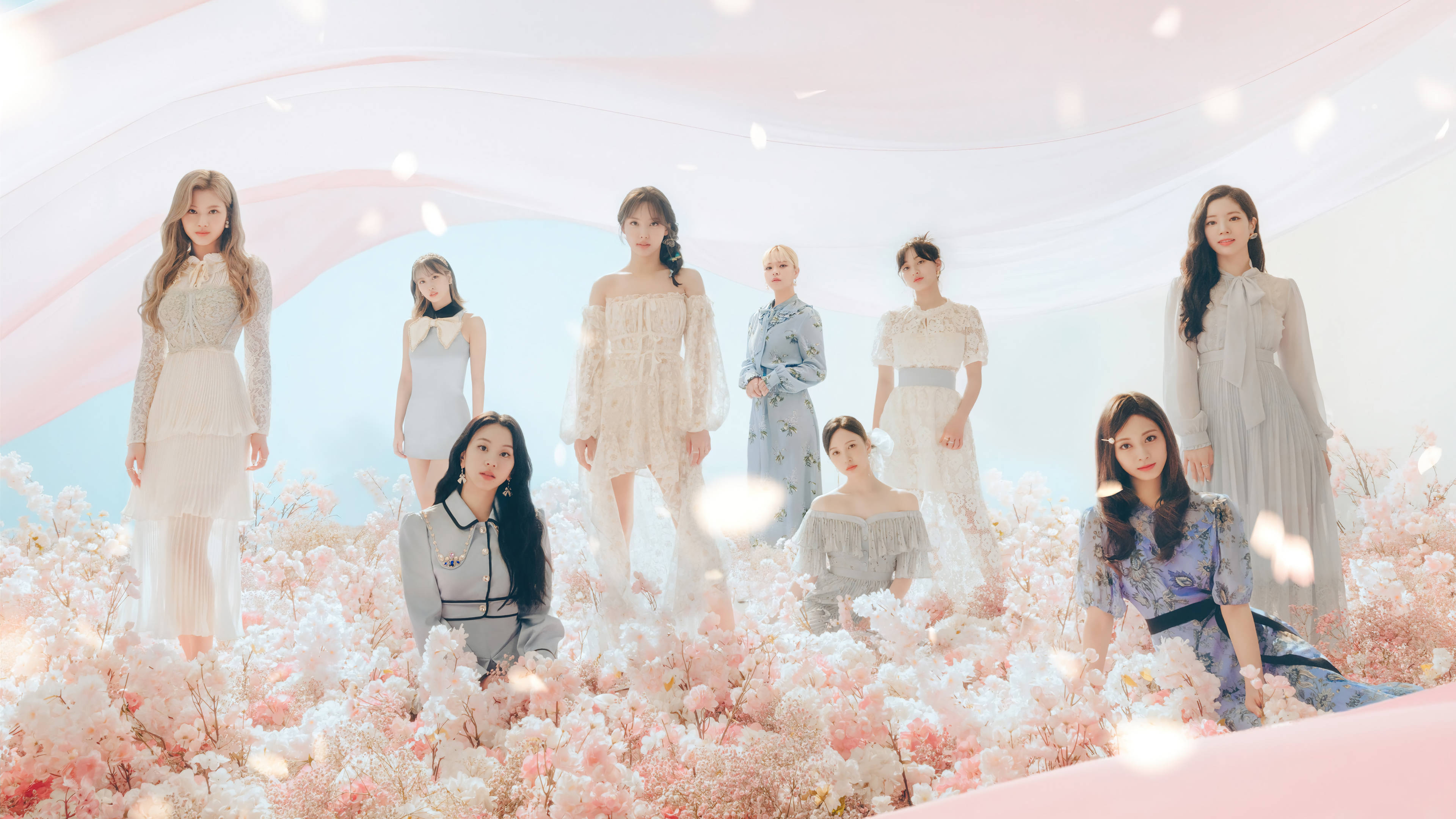 Kpop Aesthetic 桌布 壁紙 — 七位身穿優雅連衣裙的女性在夢幻般的柔和粉紅色和白色花朵中優雅地擺姿勢，背景是夢幻般的粉彩天空。