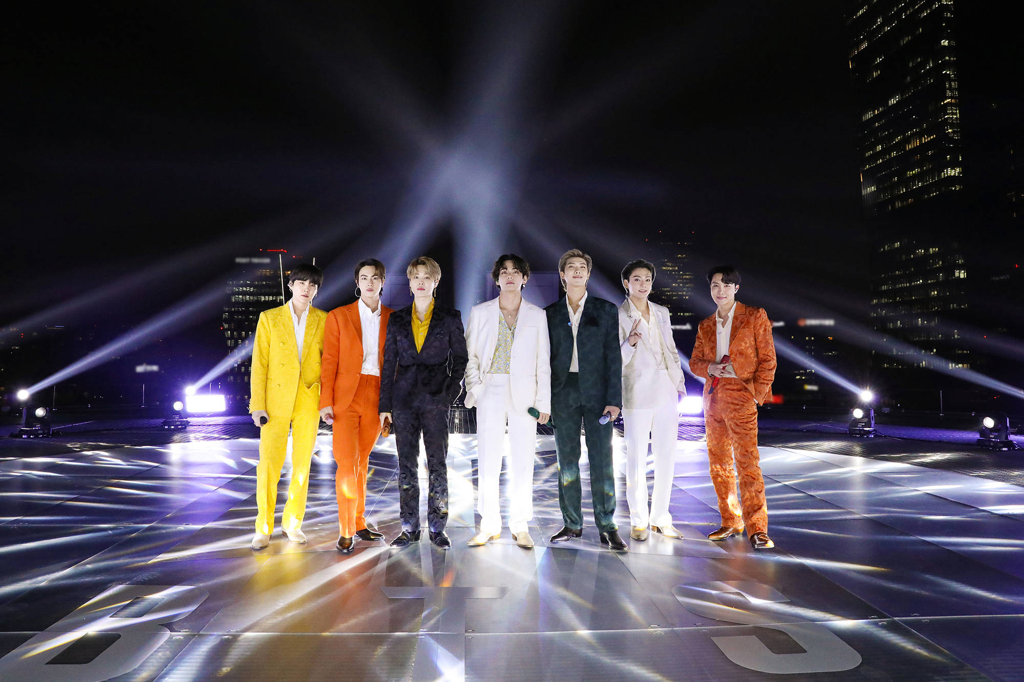 BTS 2021 桌布 壁紙 — 七位身穿大膽色彩西裝的男士，在夜間城市天際線的背景下，自信地站在戲劇性聚光燈下的發光舞台上。