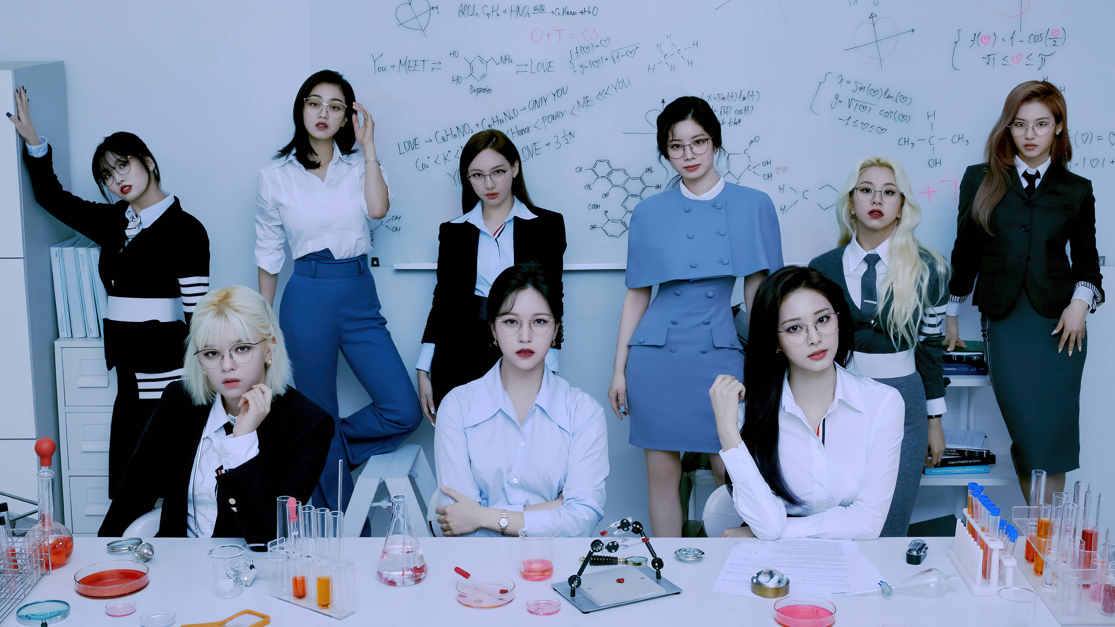 Kpop Aesthetic 桌布 壁紙 — 七位身穿實驗室服裝的女性自信地圍繞著一張放有燒杯和寫滿方程式的白板的科學桌擺姿勢。