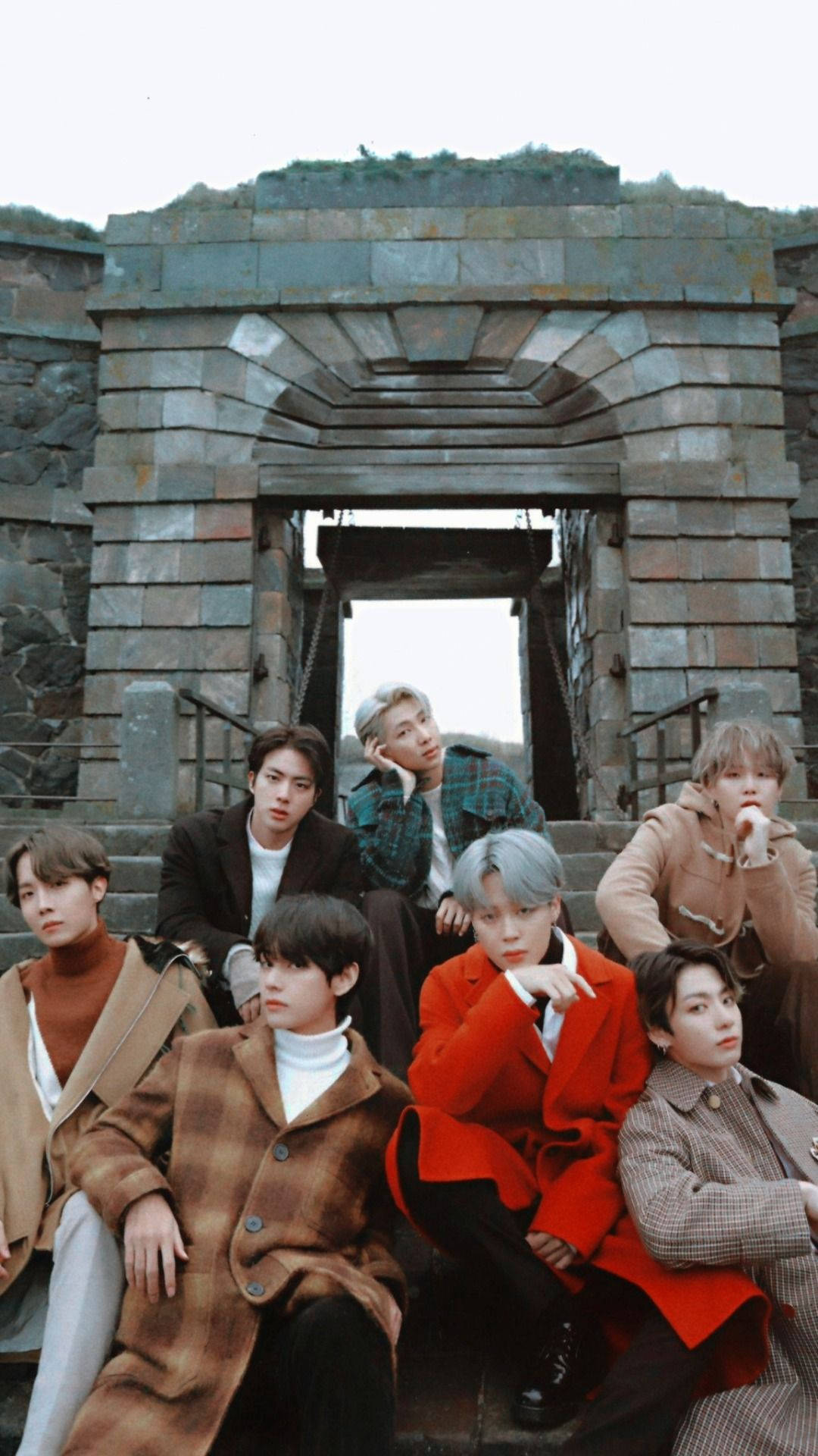 BTS 桌布 壁紙 — 七位身穿時尚外套的年輕男子在風化的石拱門下的石階上擺姿勢，背景是柔和天空。