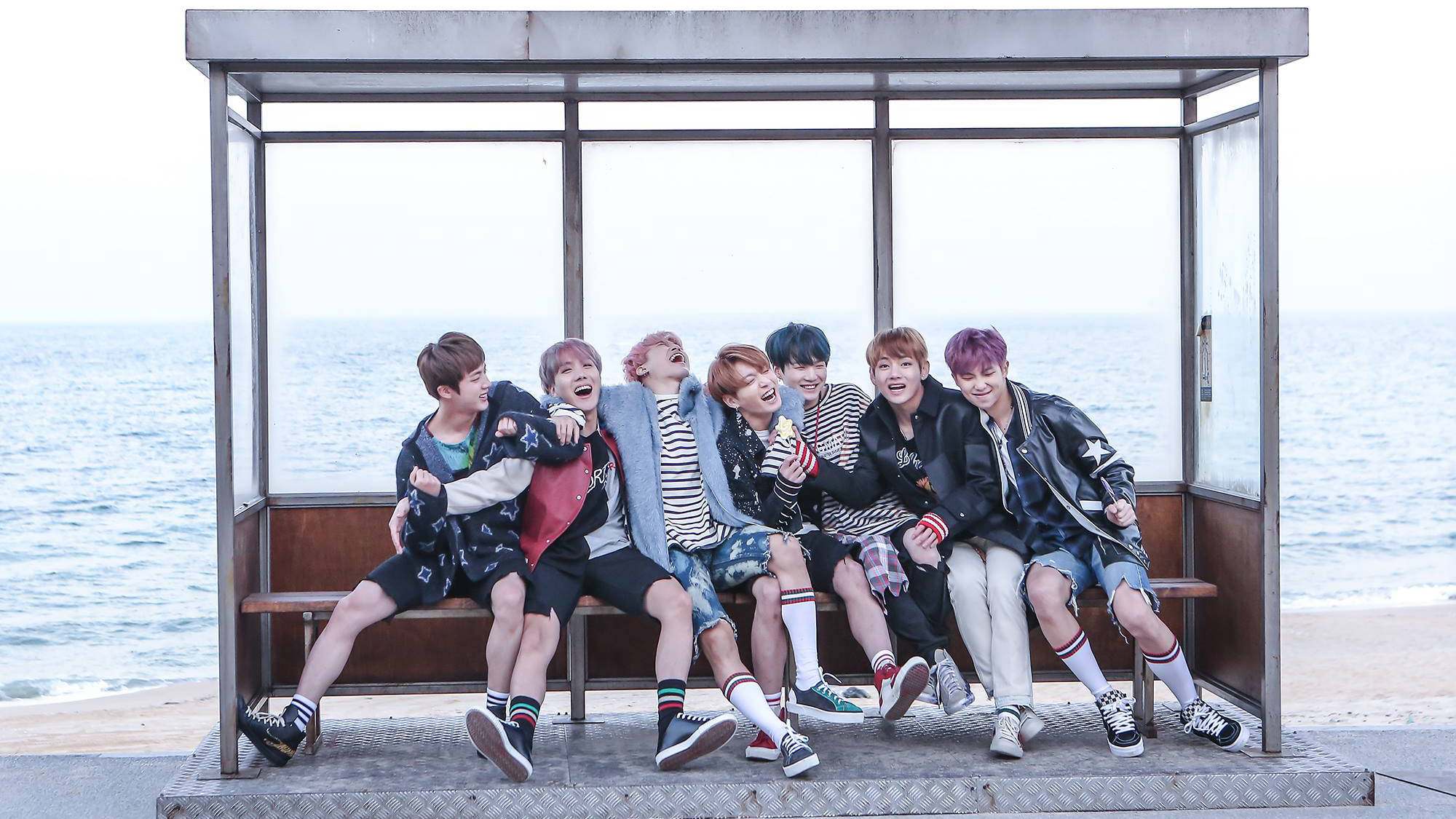 BTS 桌布 壁紙 — 七位身穿時尚街頭服的年輕男士在海邊巴士站長椅上大笑並擺姿勢，身後是海洋。