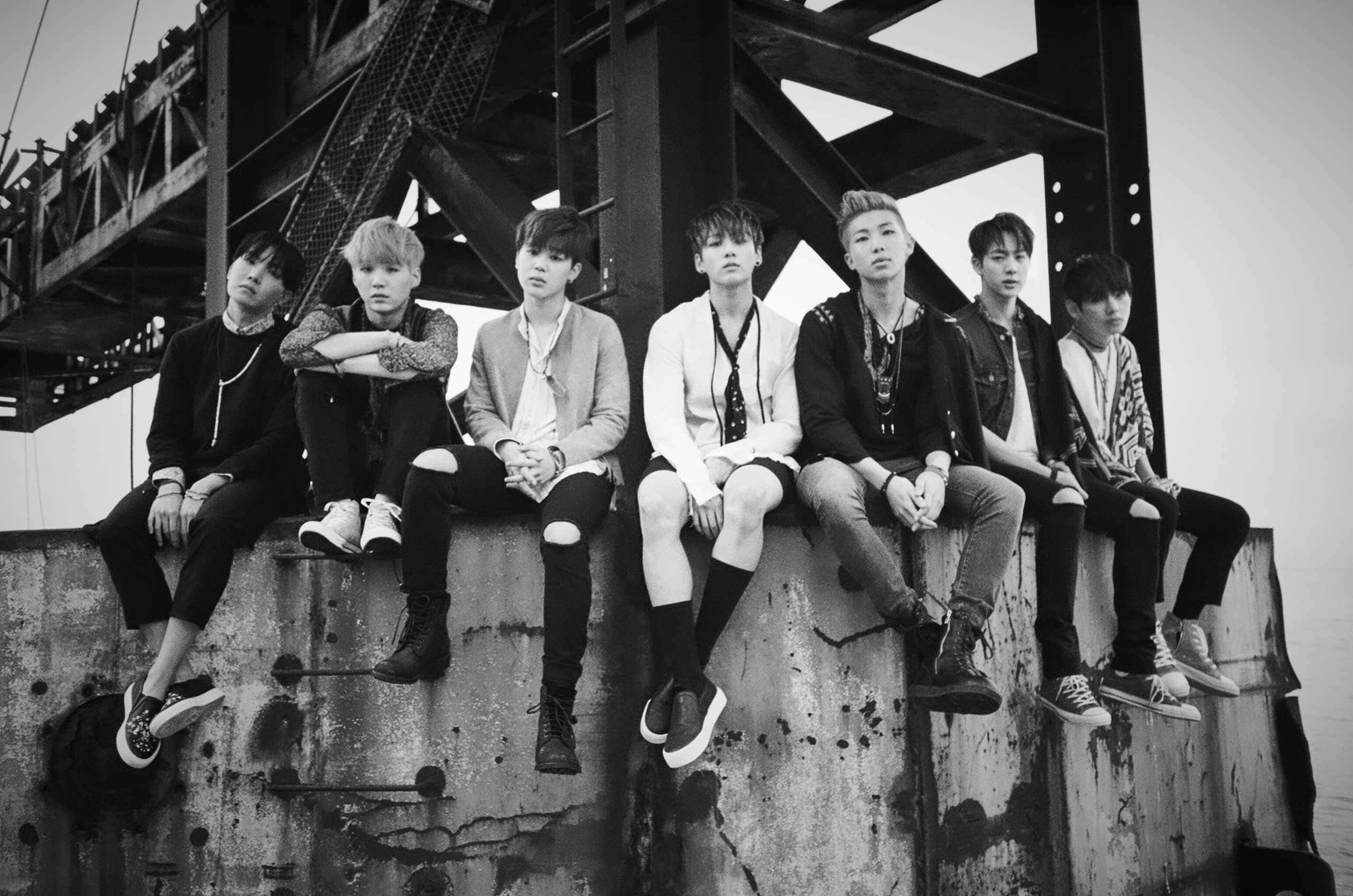 bts photoshoot 桌布 壁紙 — 七位身穿時尚街頭服的年輕男子，在巨型鋼橋下，隨意坐在風化的混凝土邊緣，以黑白呈現。