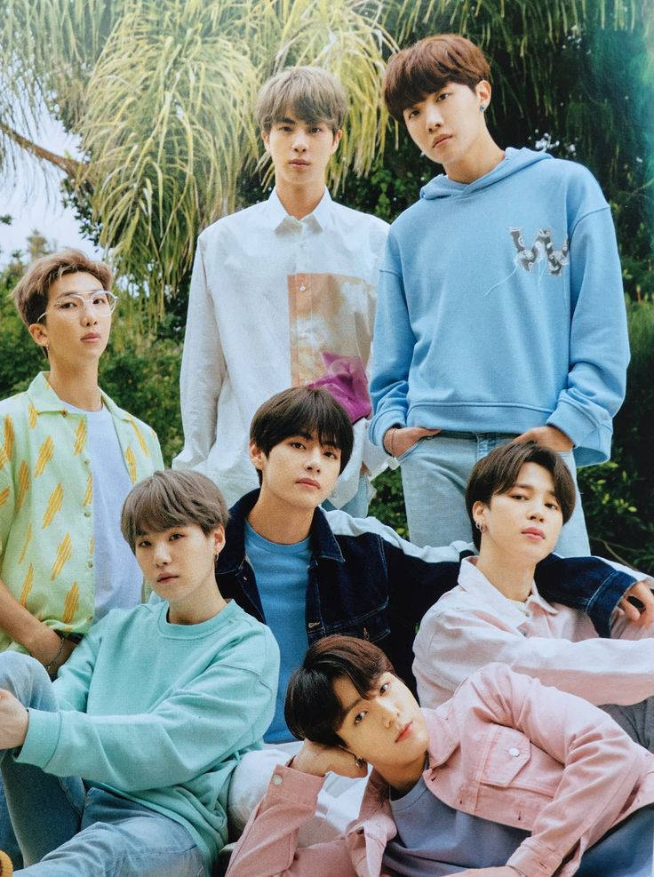 BTS 桌布 壁紙 — 七位身穿柔和色調休閒服的年輕男子在明亮的日光下，自信地站在茂密的綠色棕櫚樹中擺姿勢。