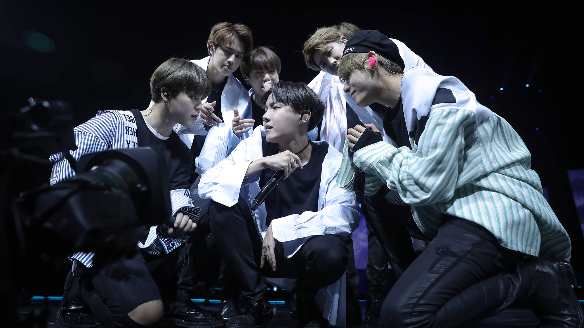 bts group cute 桌布 壁紙 — 七位身穿條紋襯衫的年輕男子在演唱會期間，於明亮的聚光燈下緊密地擠在黑暗的舞台上。