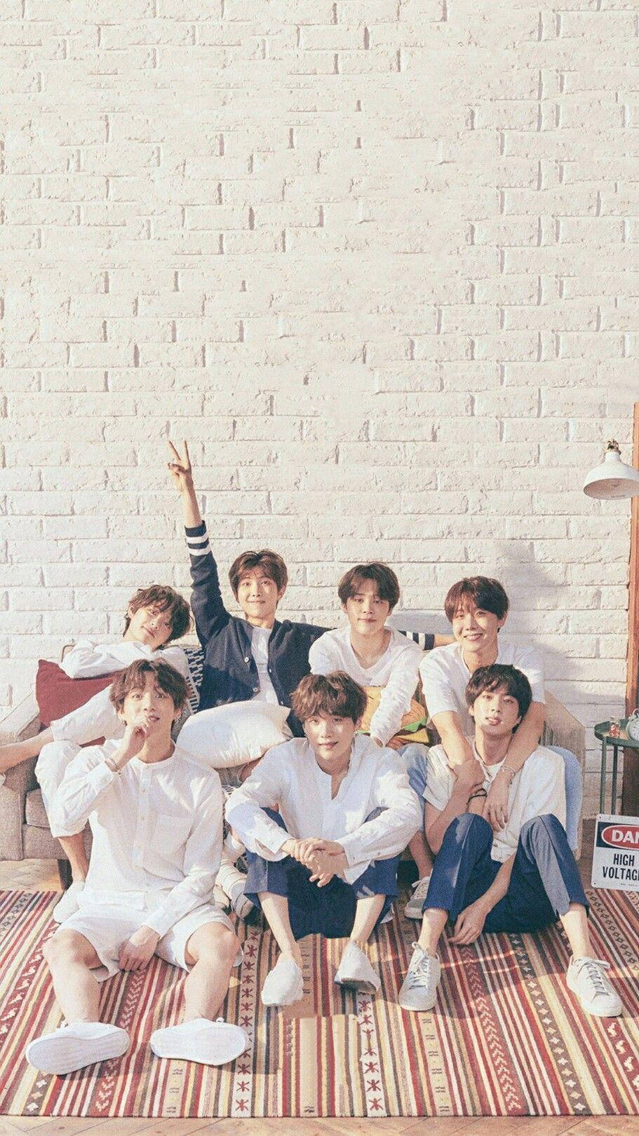 BTS 桌布 壁紙 — 七位身穿白色休閒裝的年輕男子在圖案地毯上，以白色磚牆為背景，愉快地擺姿勢。