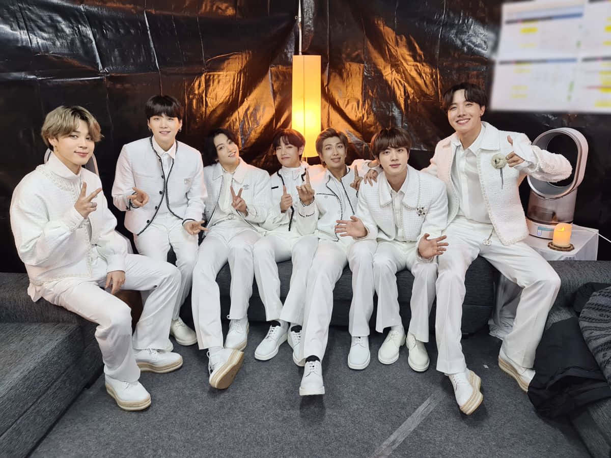 Big Hit Entertainment 桌布 壁紙 — 七位身穿白色套裝的男子在黑色背景和溫暖燈光下，於灰色沙發上愉快地擺姿勢。