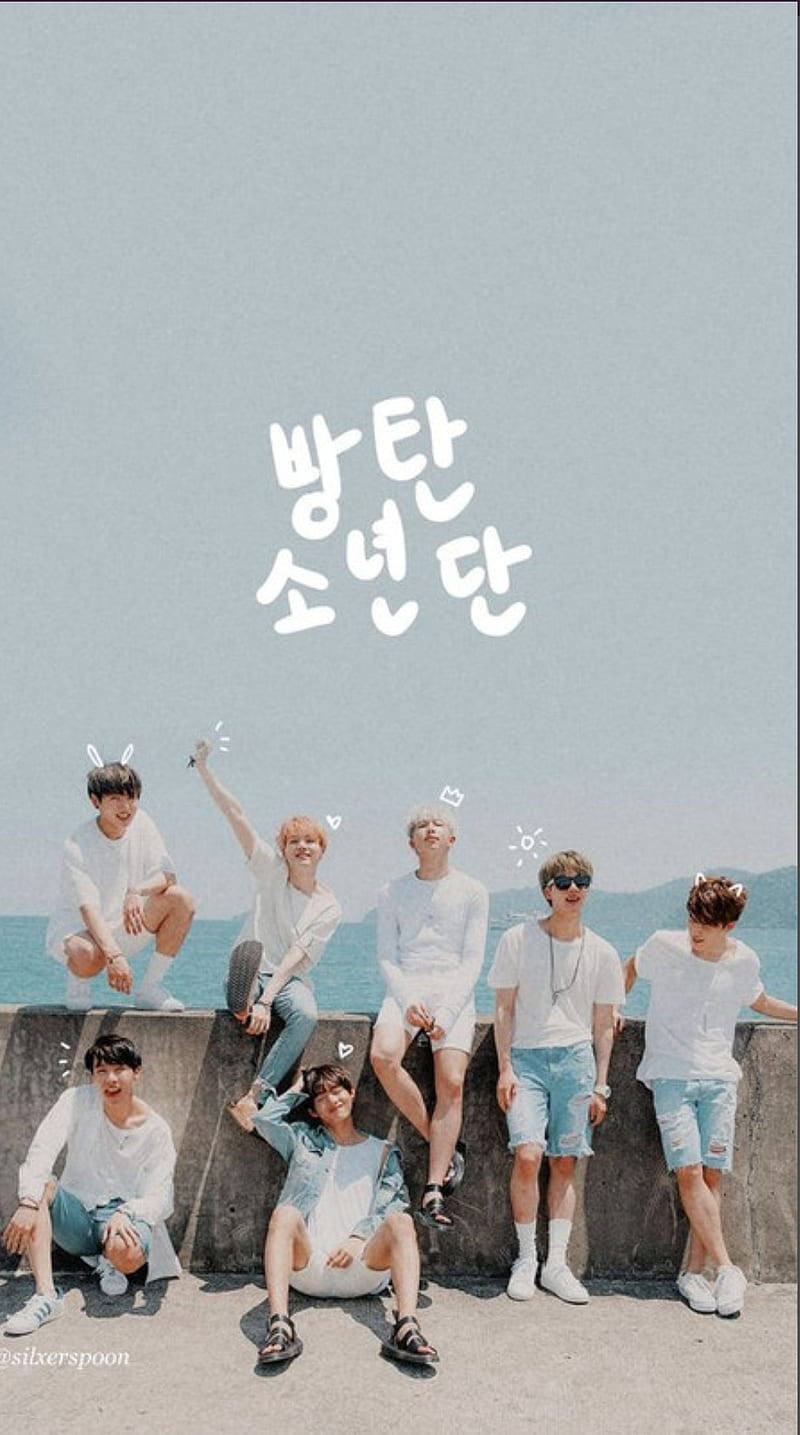 cute bts group 桌布 壁紙 — 七位身穿白色服裝的年輕男子在淡藍色天空下的海邊混凝土牆上愉快地擺姿勢，上方漂浮著韓文文字。