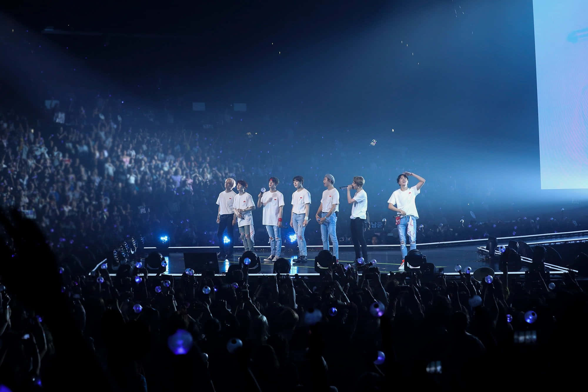 Bts Concert 桌布 壁紙 — 七位身穿白襯衫的表演者站在燈光明亮的舞台上，面對著一大群歡呼的觀眾，他們手持發光的手機。