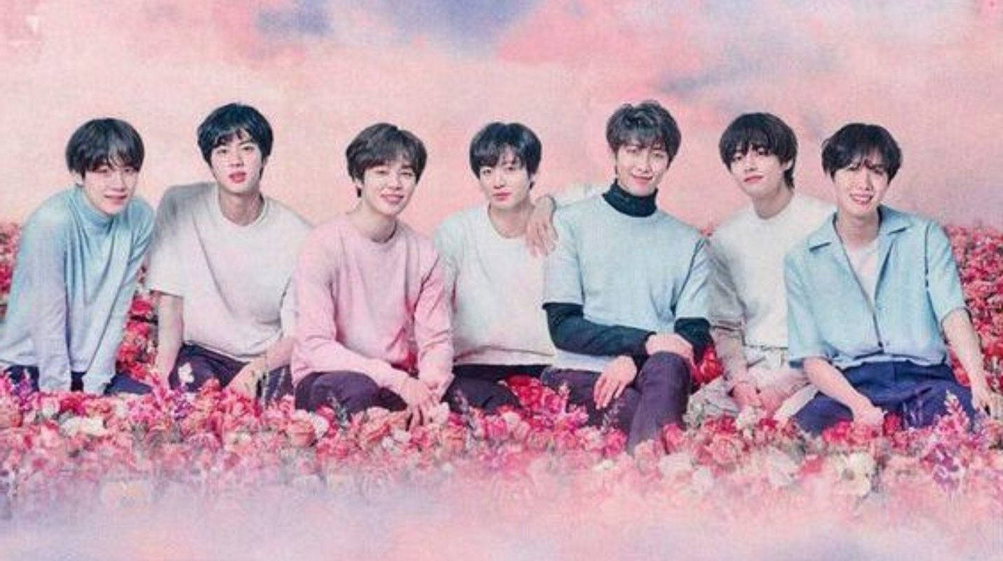 cute bts group 桌布 壁紙 — 七位身穿粉彩服裝的年輕男子，在夢幻般的粉紅天空下，微笑著坐在一片粉紅玫瑰海中。