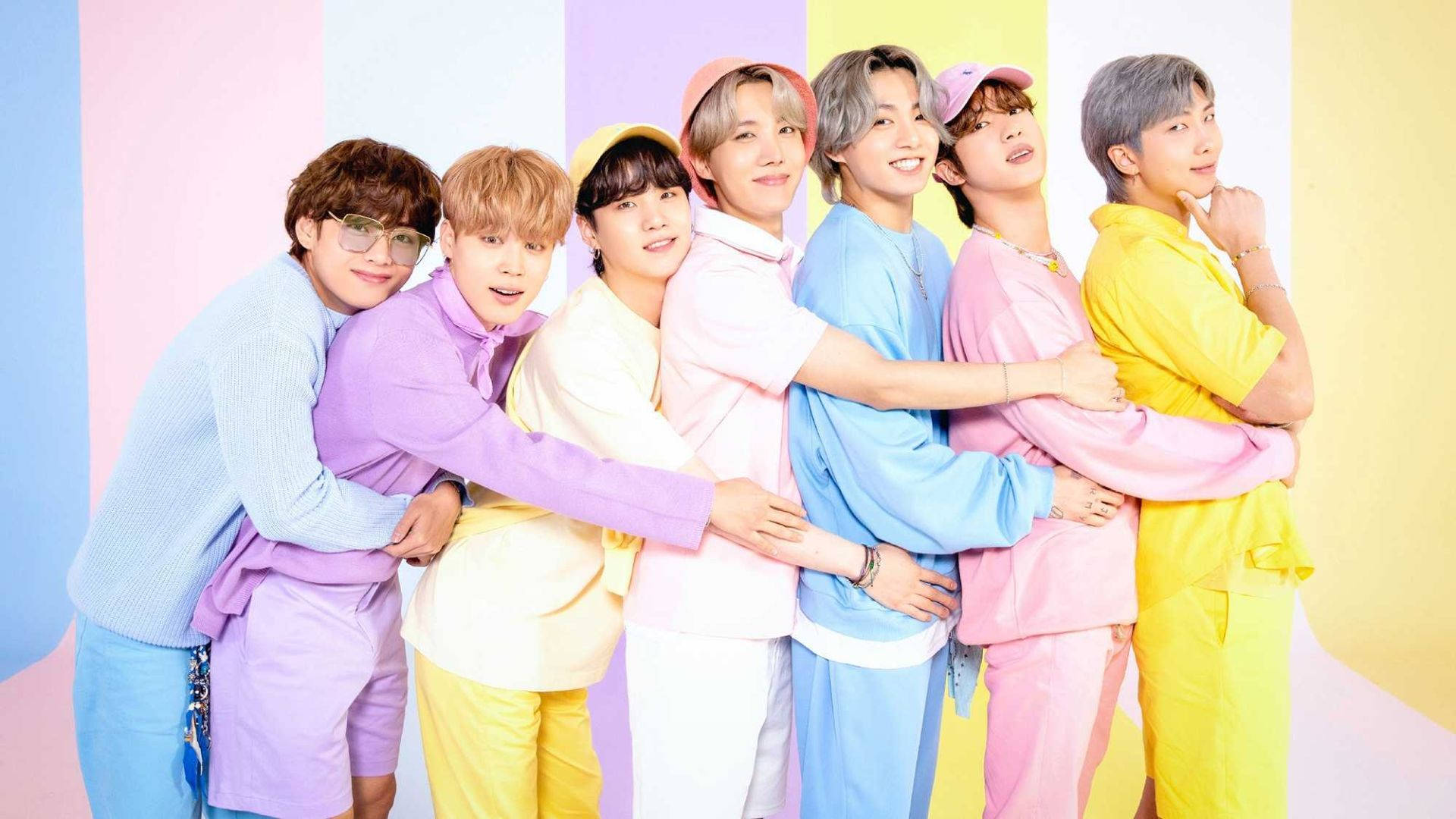 BTS 桌布 桌布 壁紙 — 七位身穿粉彩服裝的年輕男子，在色彩繽紛的條紋背景前，面帶愉快表情互相挽著手臂。