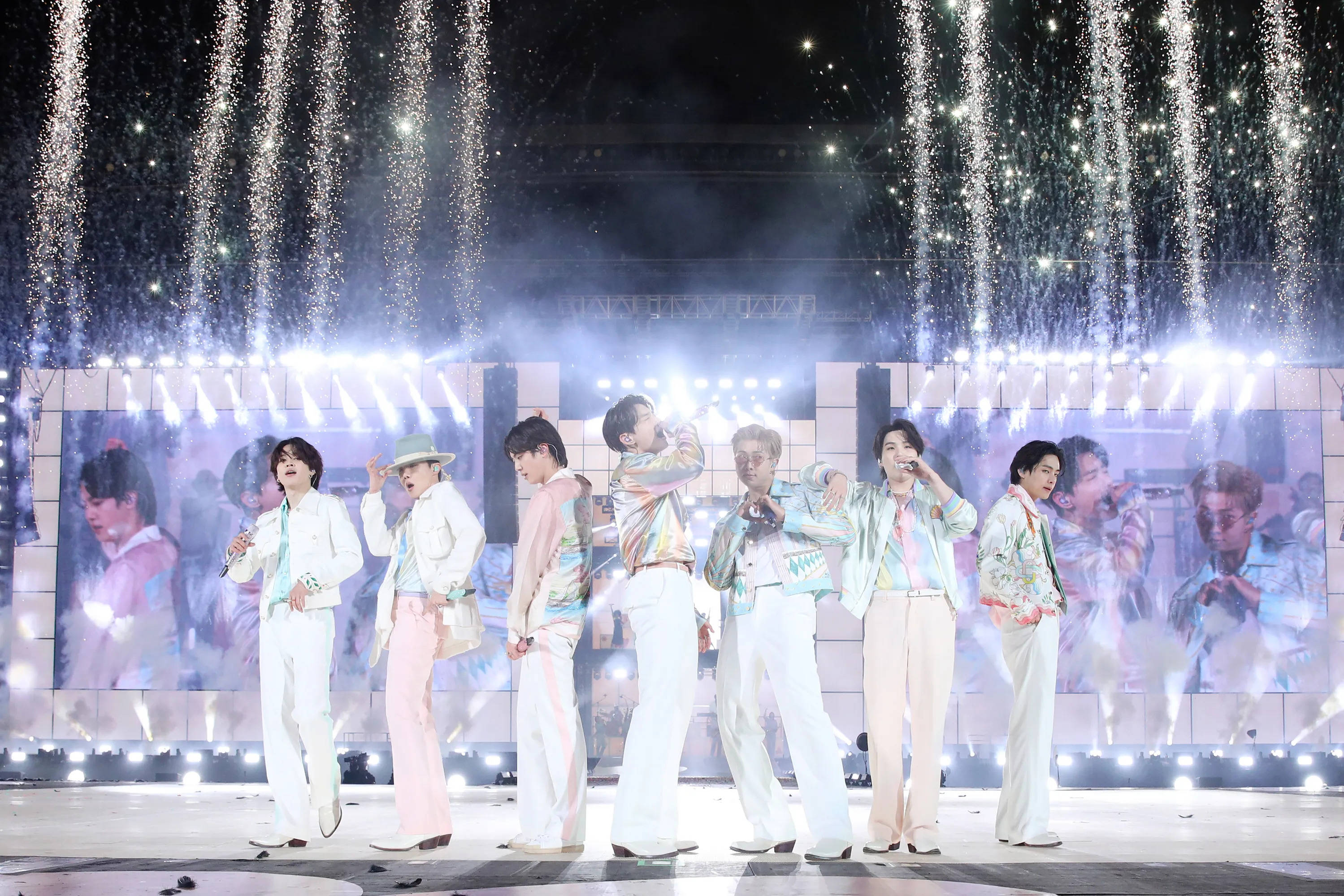 Bts Aesthetic 桌布 壁紙 — 七位身穿粉彩服裝的表演者在閃爍的舞台燈光和飄落的亮片下於音樂會舞台上跳舞。