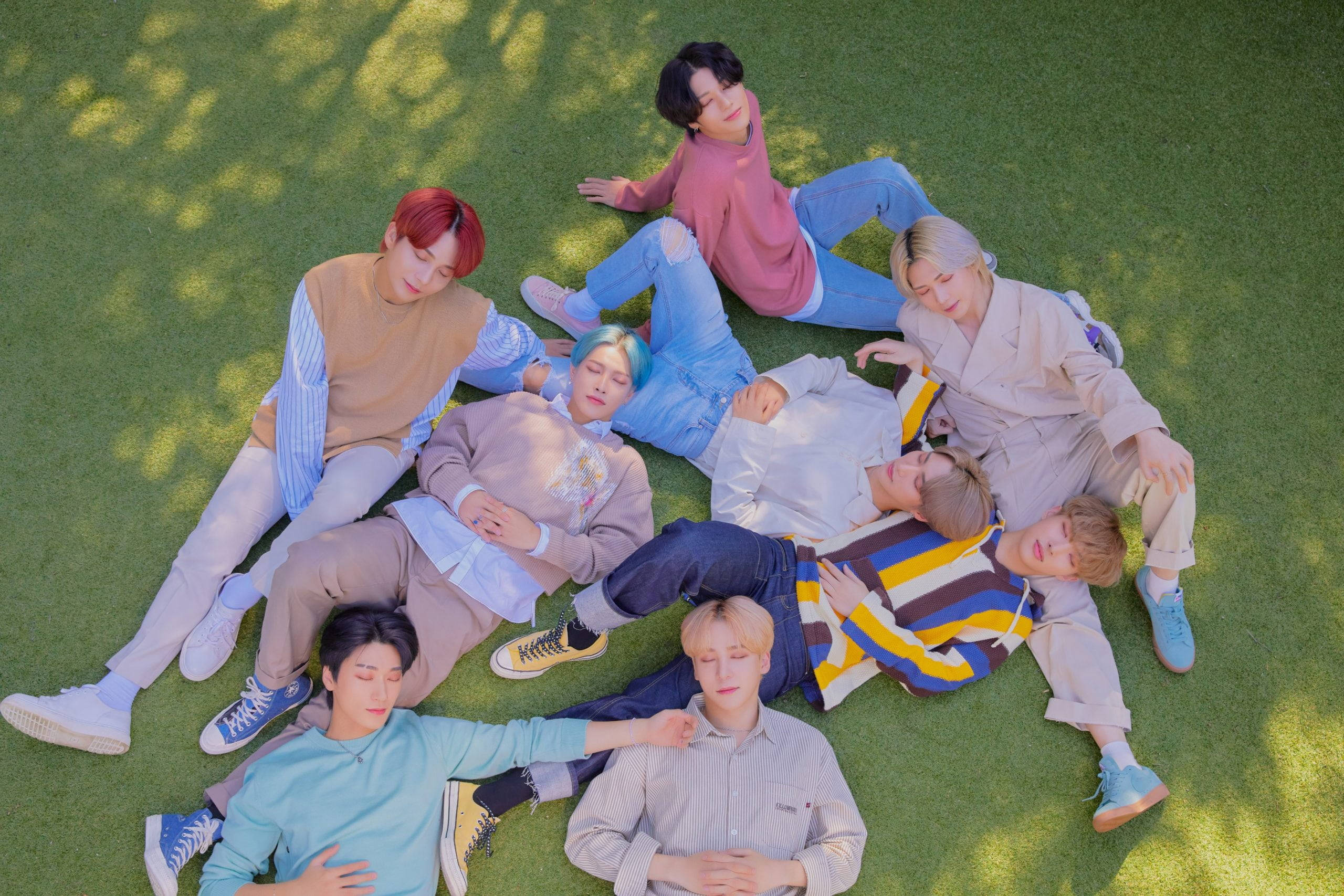ateez 桌布 壁紙 — 七位身穿色彩繽紛休閒服的年輕男子，在斑駁的陽光下圍坐在鮮綠色的草地上。