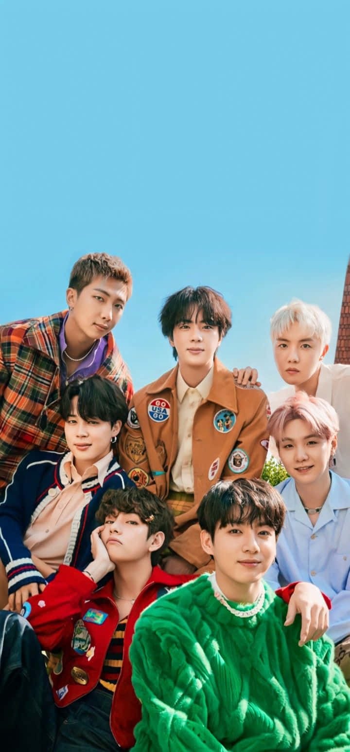 BTS 桌布 壁紙 — 七位身穿色彩繽紛復古服裝的年輕男子，在晴朗的藍天下，以磚砌建築為背景一起擺姿勢。