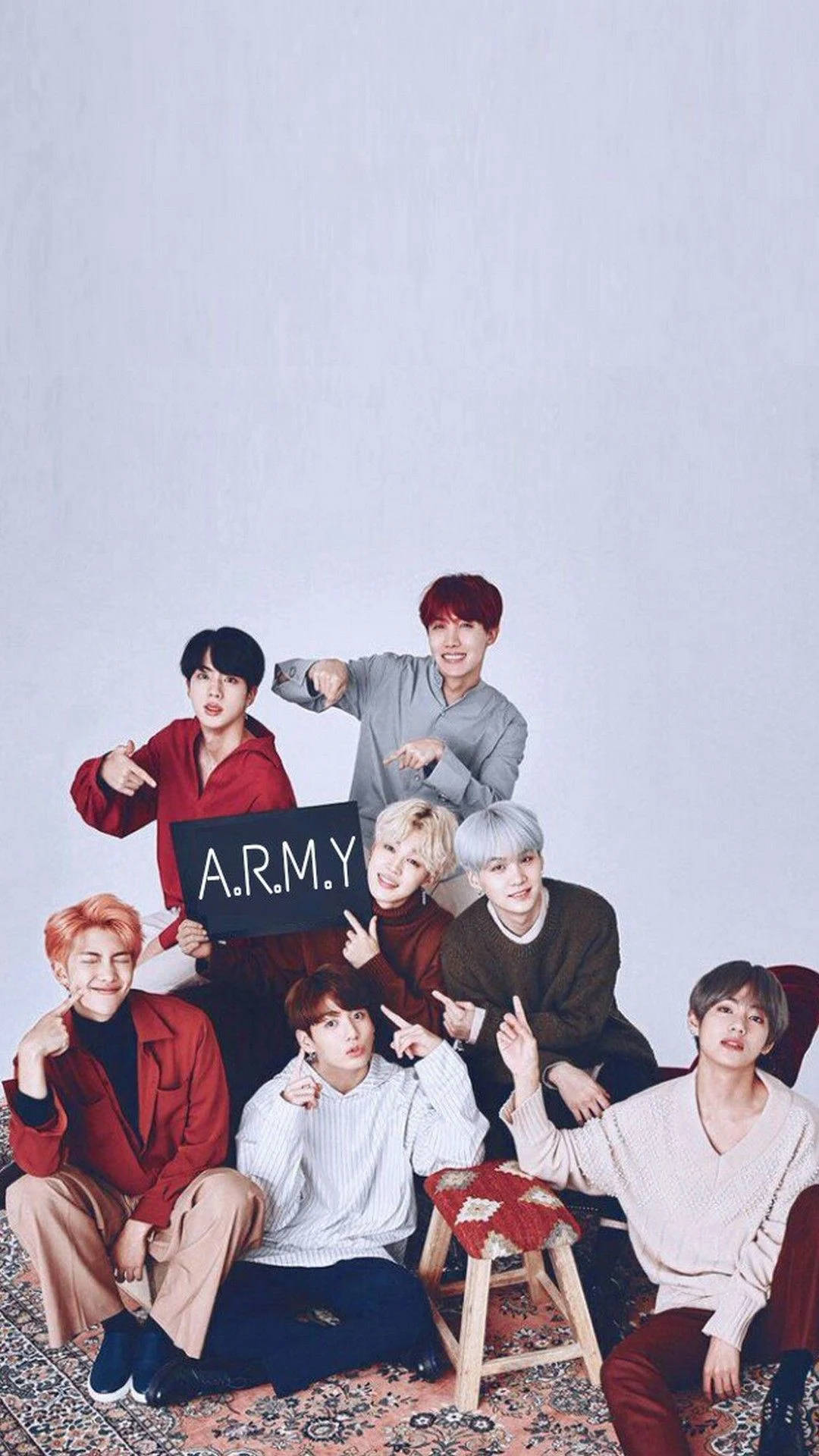 bts group cute 桌布 壁紙 — 七位身穿色彩繽紛服裝的年輕男子，在有圖案的地毯上圍繞著寫有A.R.M.Y的標誌牌擺出俏皮的姿勢，背景是素牆。