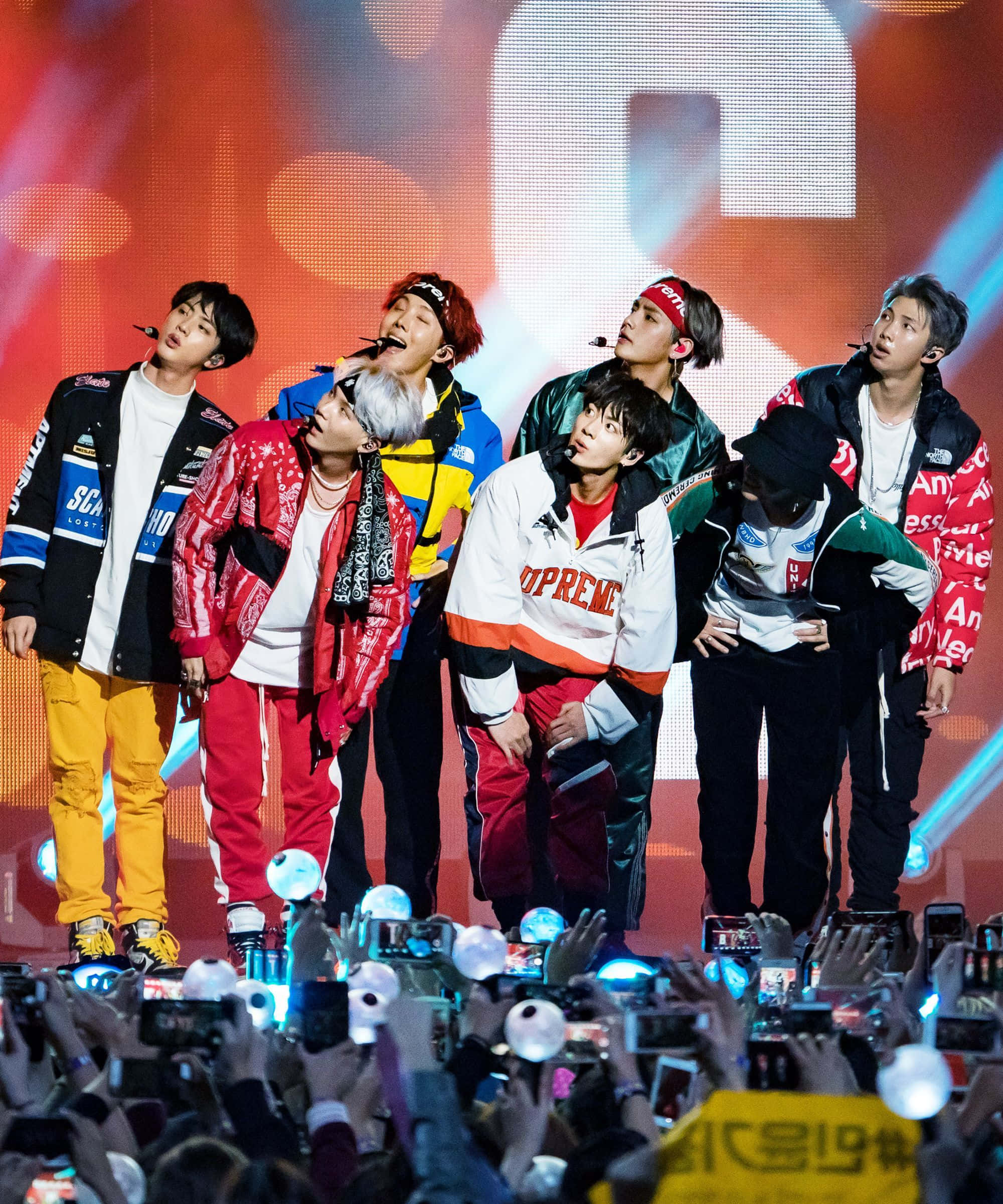 Bts Live 桌布 壁紙 — 七位身穿色彩繽紛街頭服的男K-pop偶像，在充滿活力的舞台上，伴隨著閃爍燈光和下方歡呼的人群，充滿活力地表演。