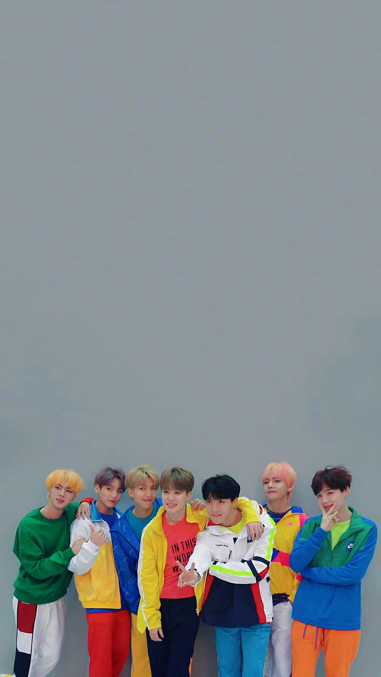 BTS 桌布 壁紙 — 七位身穿鮮豔多彩街頭服飾的年輕男性，在樸素的灰色牆壁前帶著愉快表情一起擺姿勢。