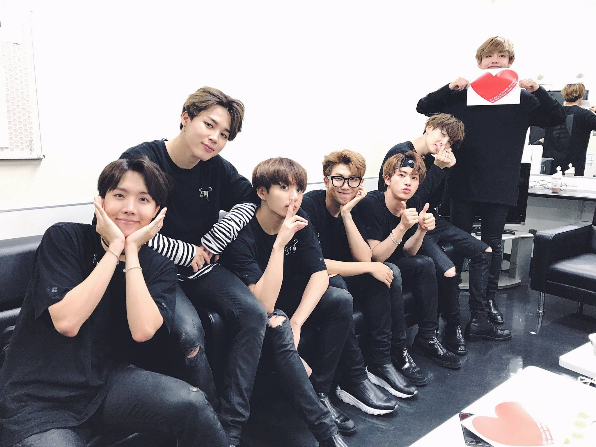 BTS 桌布 壁紙 — 七位身穿黑色服裝的年輕男子在明亮的攝影棚中，與心形道具一起在沙發上俏皮地擺姿勢。