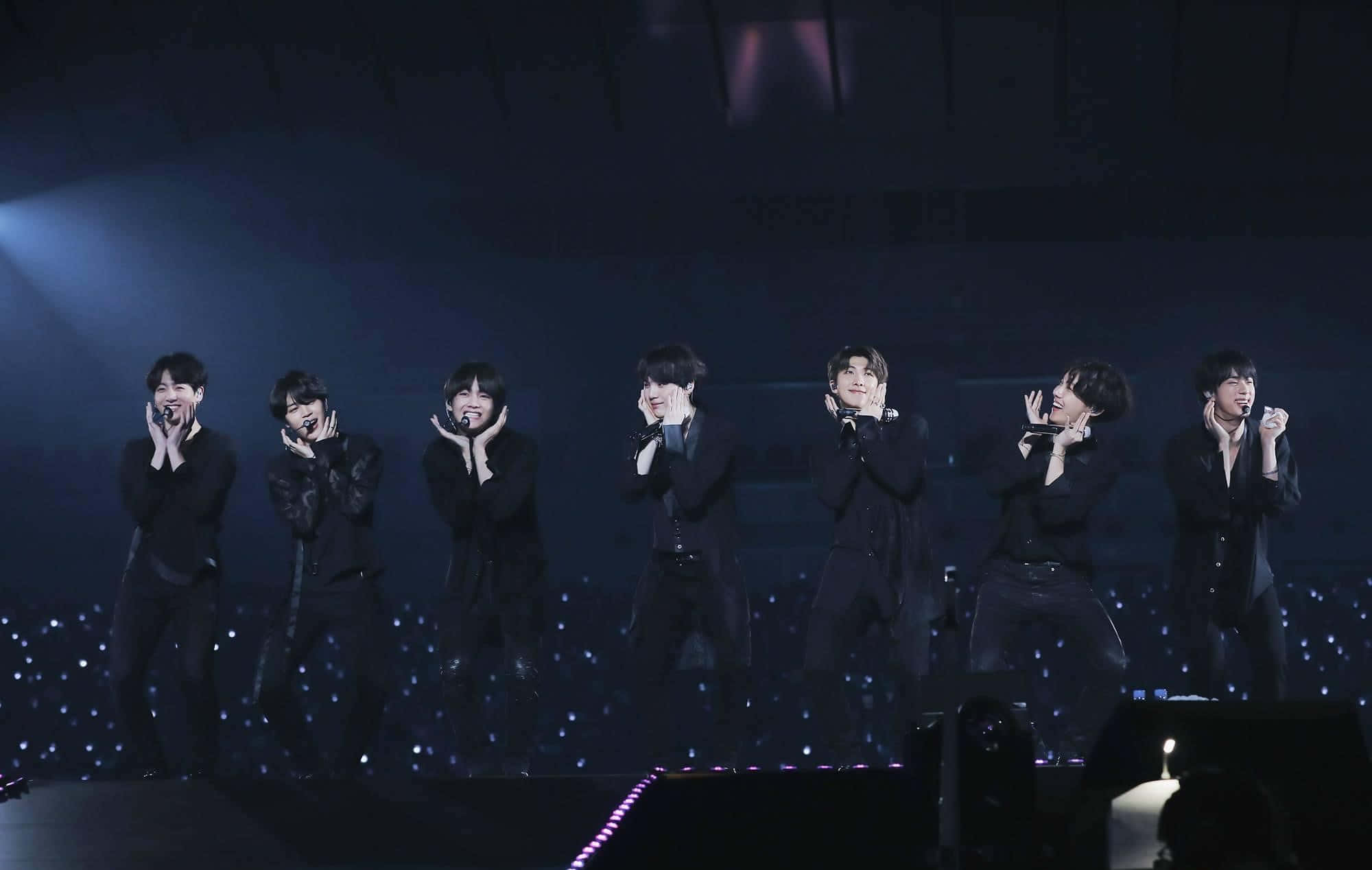 Bts Stage 桌布 壁紙 — 七位身穿黑色西裝的男表演者在藍色舞台燈光下熱情演唱，背景有發光的觀眾。