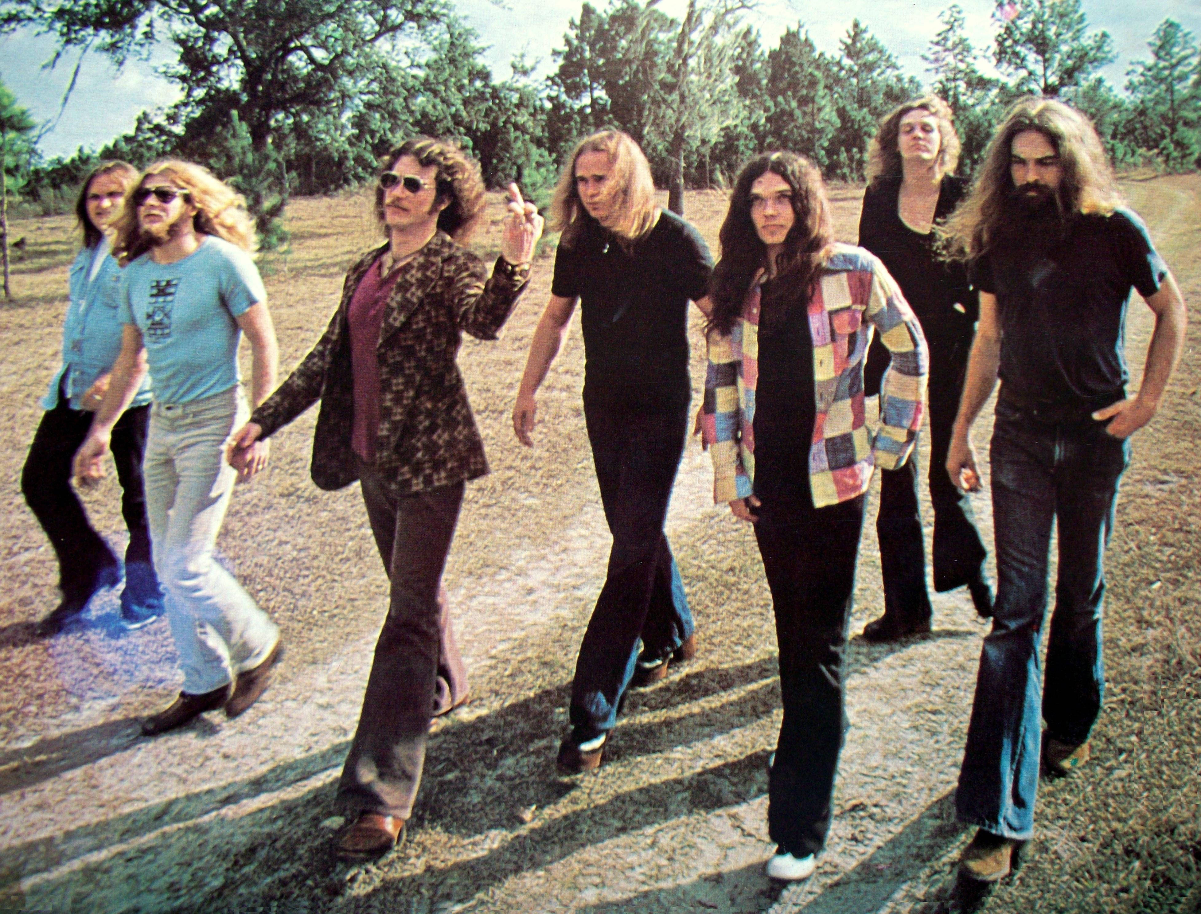 音樂 桌布 壁紙 — 七位長髮音樂家身穿1970年代服裝，自信地漫步在陽光普照的松樹林空地。