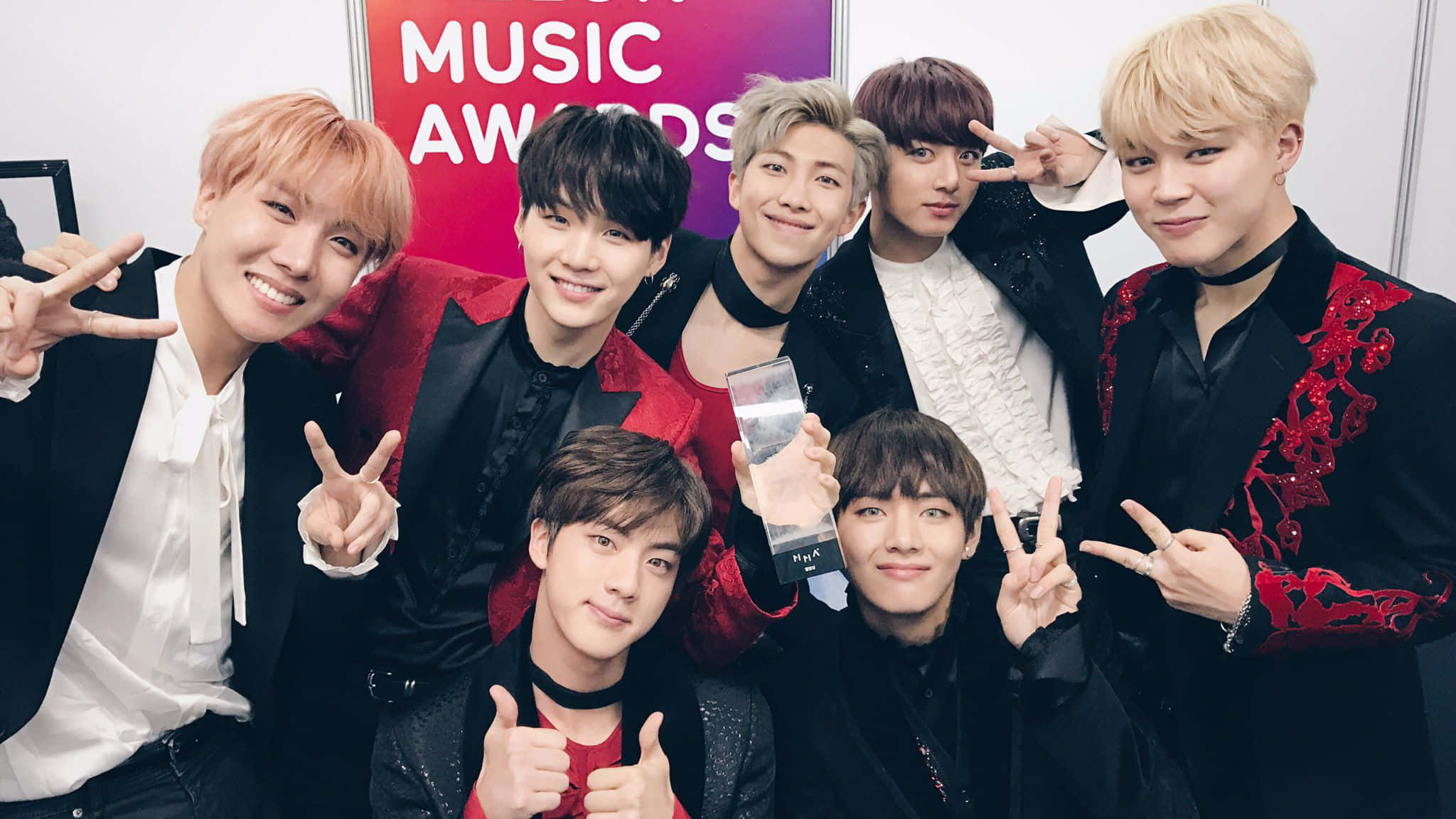 Bts Mac 桌布 壁紙 — 七位面帶微笑的K-pop偶像身穿黑色和紅色服裝，在音樂頒獎典禮上拿著獎盃擺姿勢。