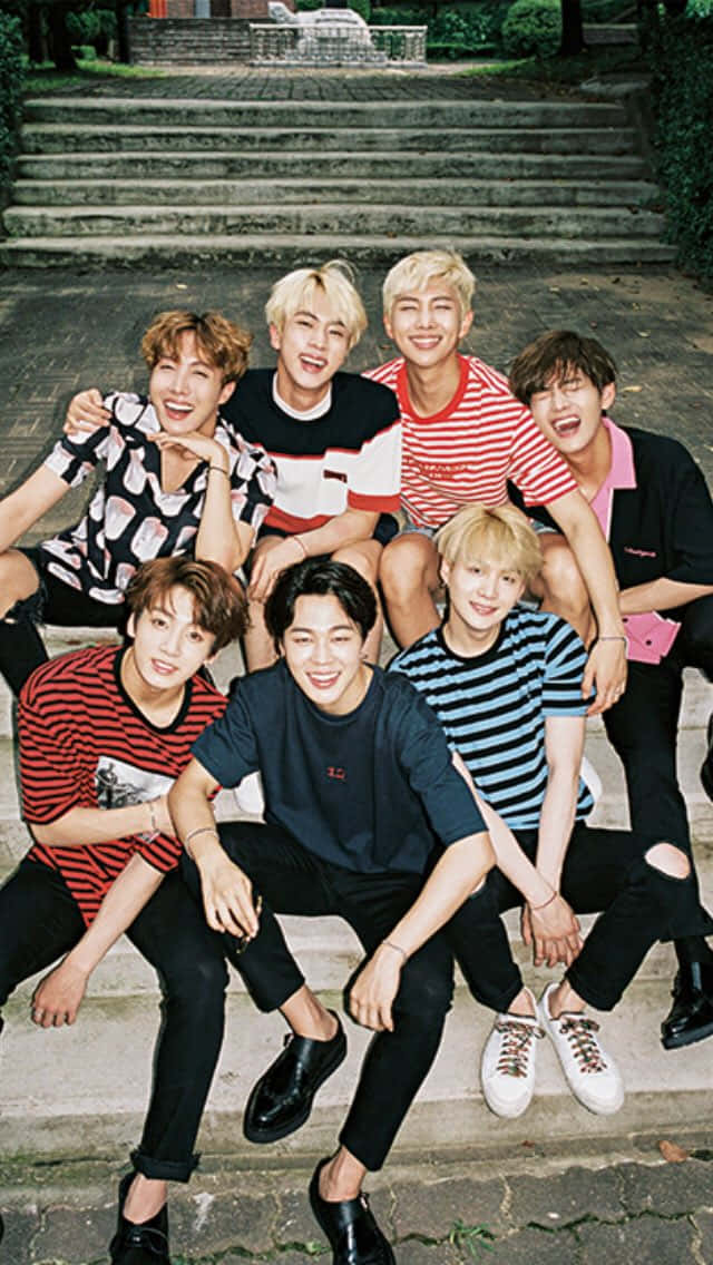 BTS 桌布 壁紙 — 七位面帶微笑的年輕男子身穿色彩繽紛的條紋襯衫，在綠意環繞的石階上愉快地擺姿勢。