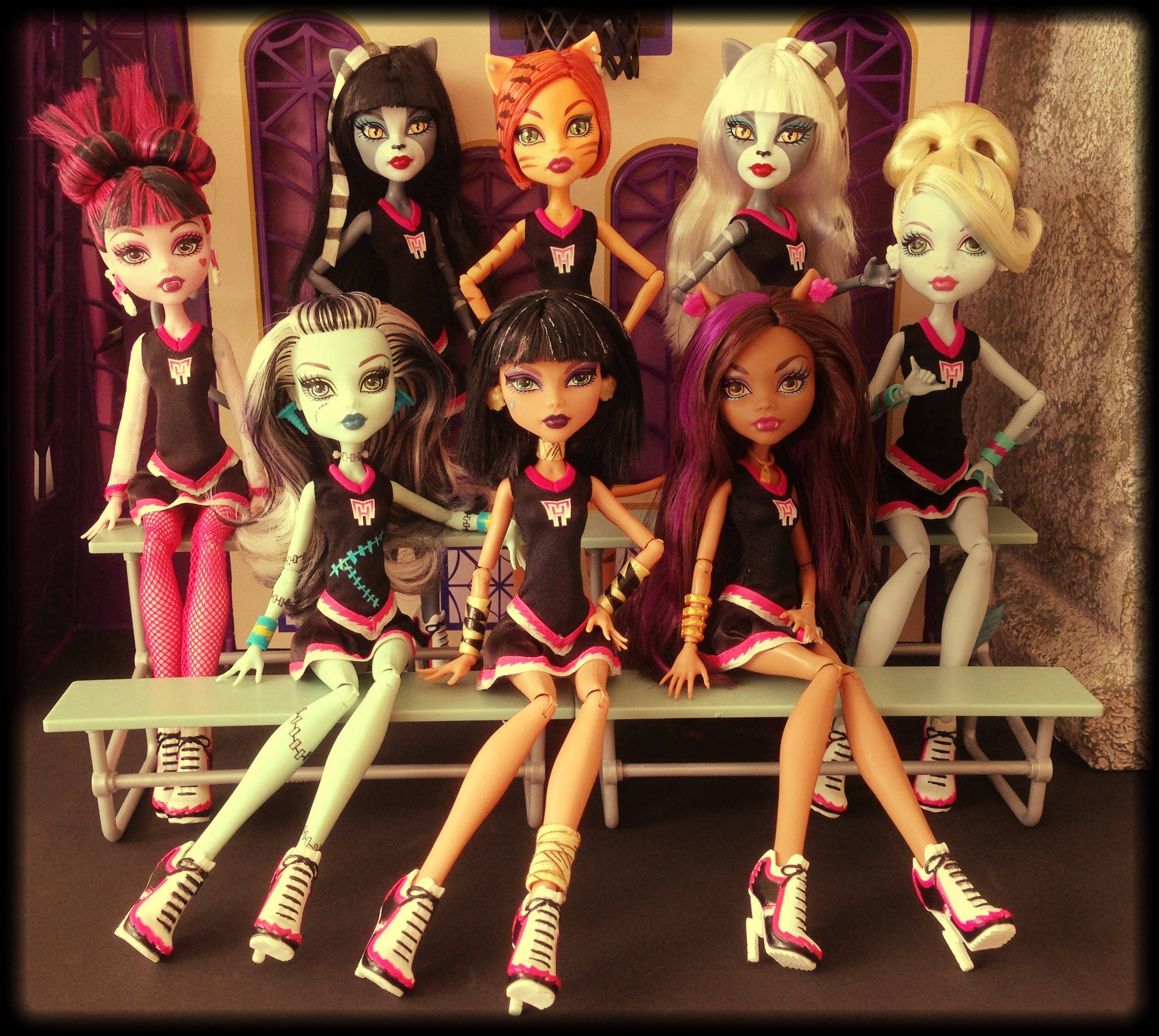 bratz 美學 桌布 壁紙 — 七個Monster High公仔穿著相配的黑色和粉紅色啦啦隊制服，在紫色拱形背景前的長椅上自信地擺姿勢。