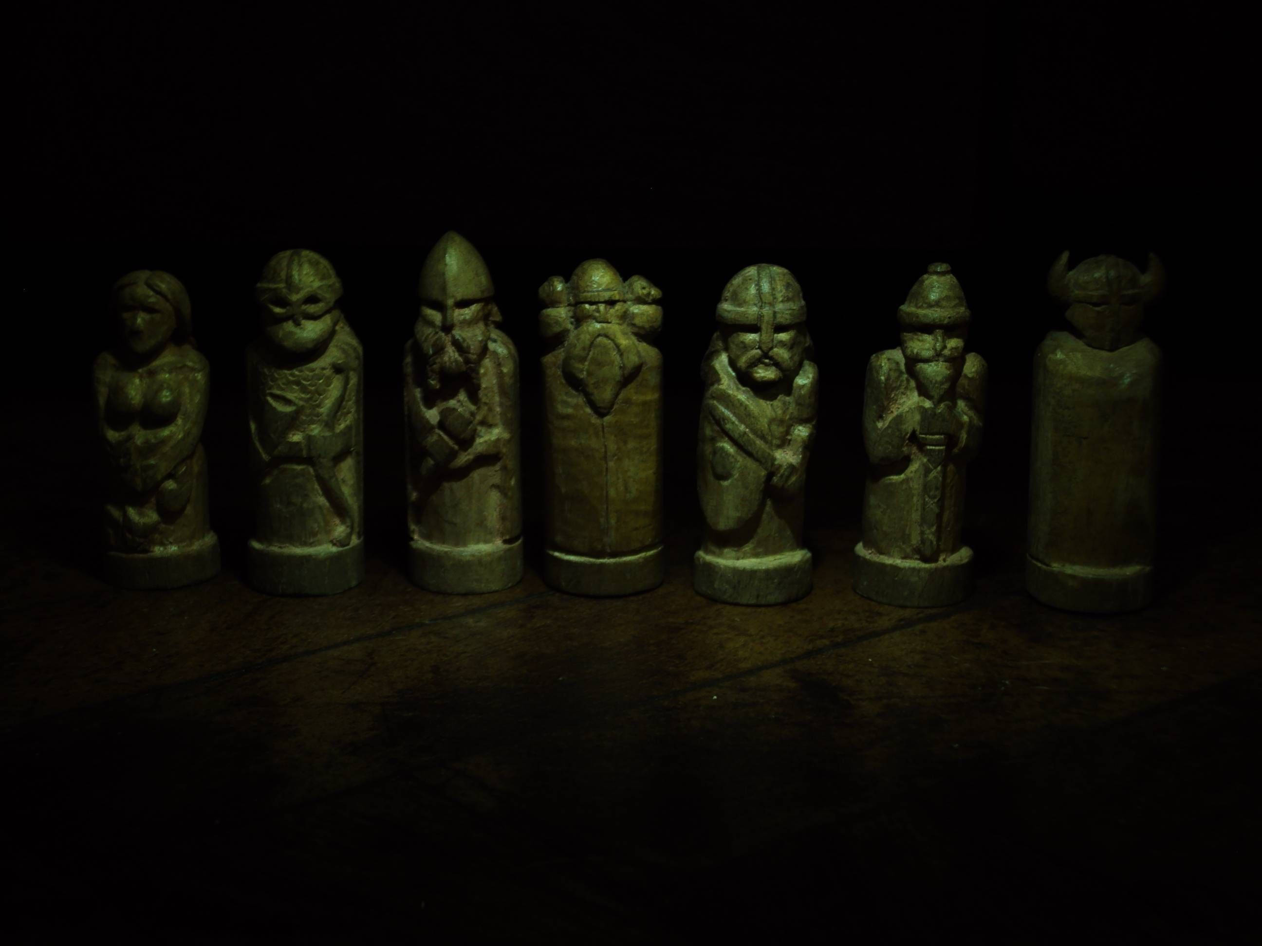 北歐 桌布 壁紙 — 七個古代雕刻的木製國際象棋棋子，在黑色背景前，昏暗的綠光下發光。
