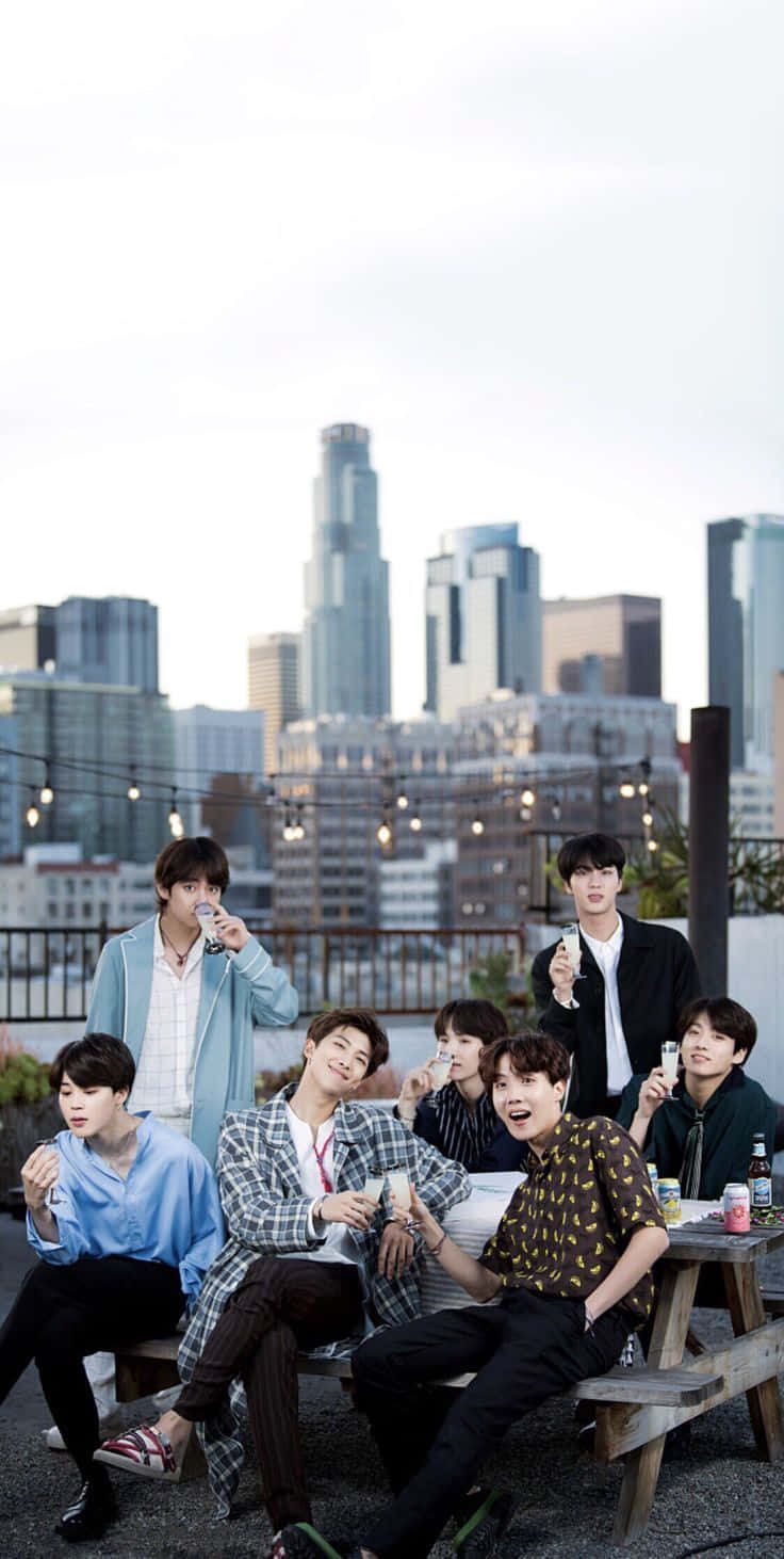 BTS 桌布 壁紙 — 七個年輕男子在屋頂野餐桌上隨意擺姿勢，背後是城市天際線，在柔和日光下。