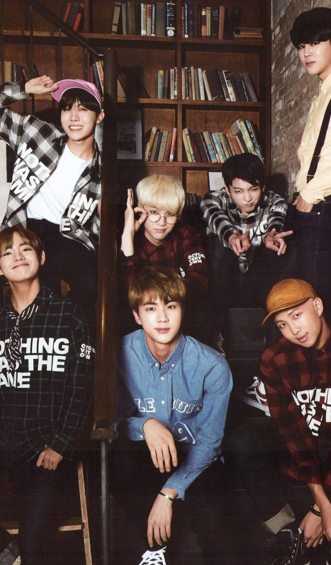 BTS 桌布 壁紙 — 七個年輕男子穿著休閒格紋和圖案T恤，在一個有木書架和磚牆的舒適圖書館裡頑皮地擺姿勢。