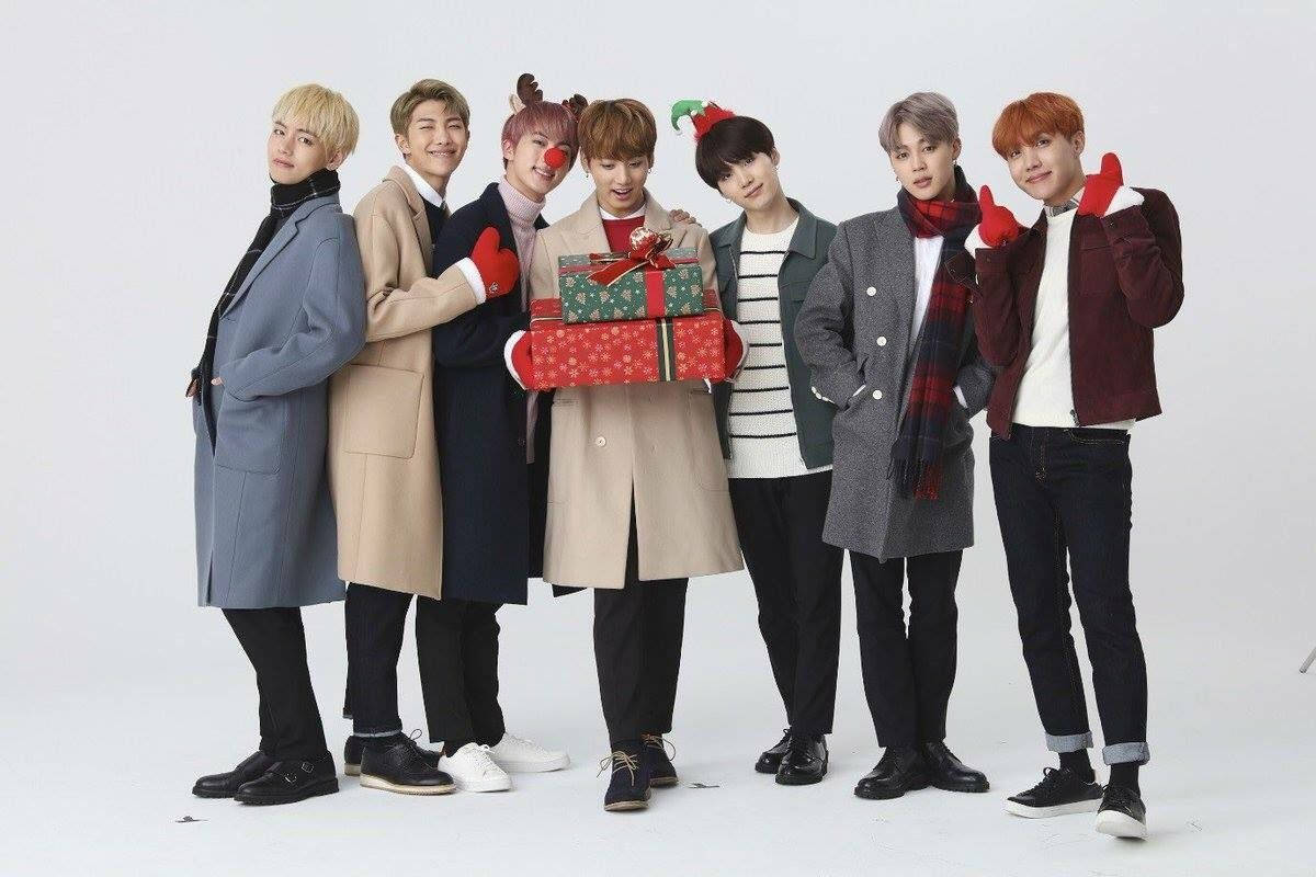 BTS 桌布 桌布 壁紙 — 七個年輕男子穿著冬季外套和節日帽子，在純白色背景前開心地拿著包裝好的禮物擺姿勢。