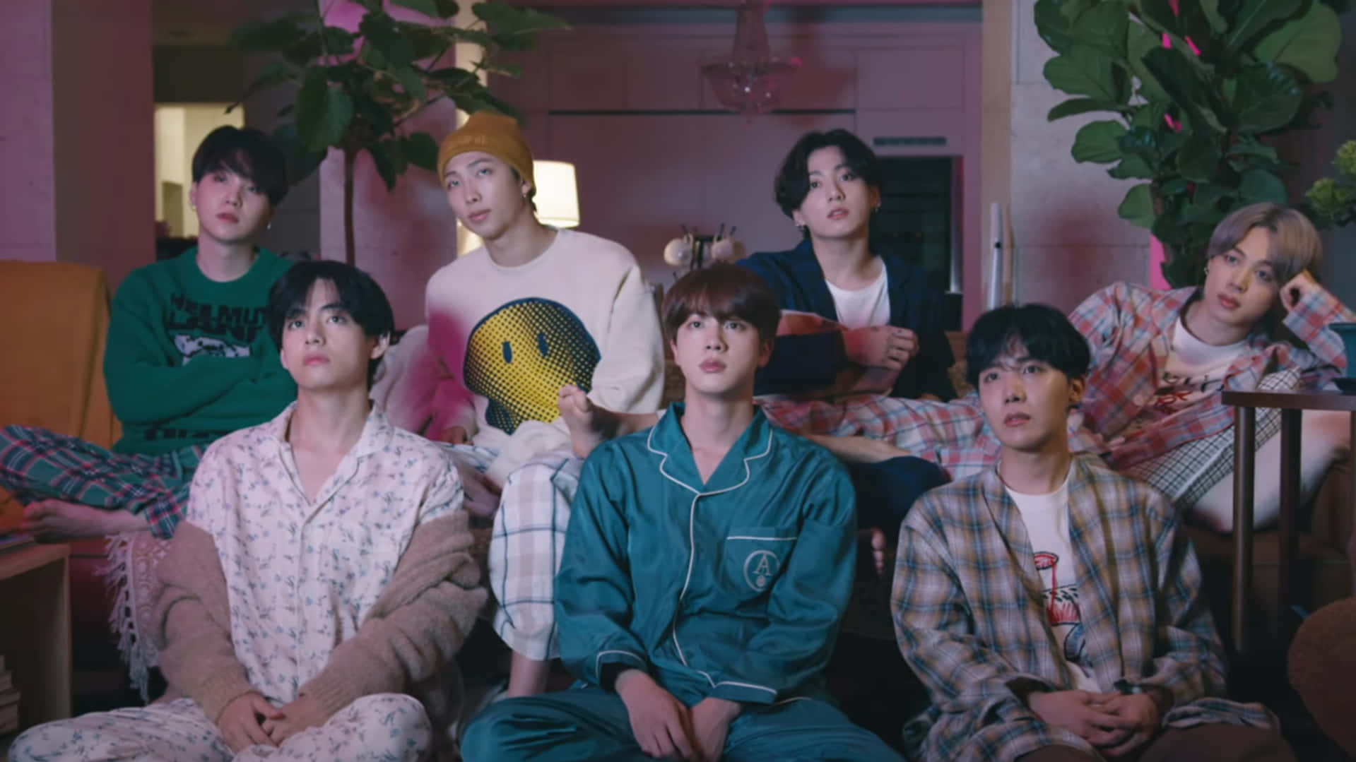 Bts Mv 桌布 壁紙 — 七個年輕男子穿著舒適睡衣和家居服，坐在一個溫暖明亮、被植物環繞的室內空間。