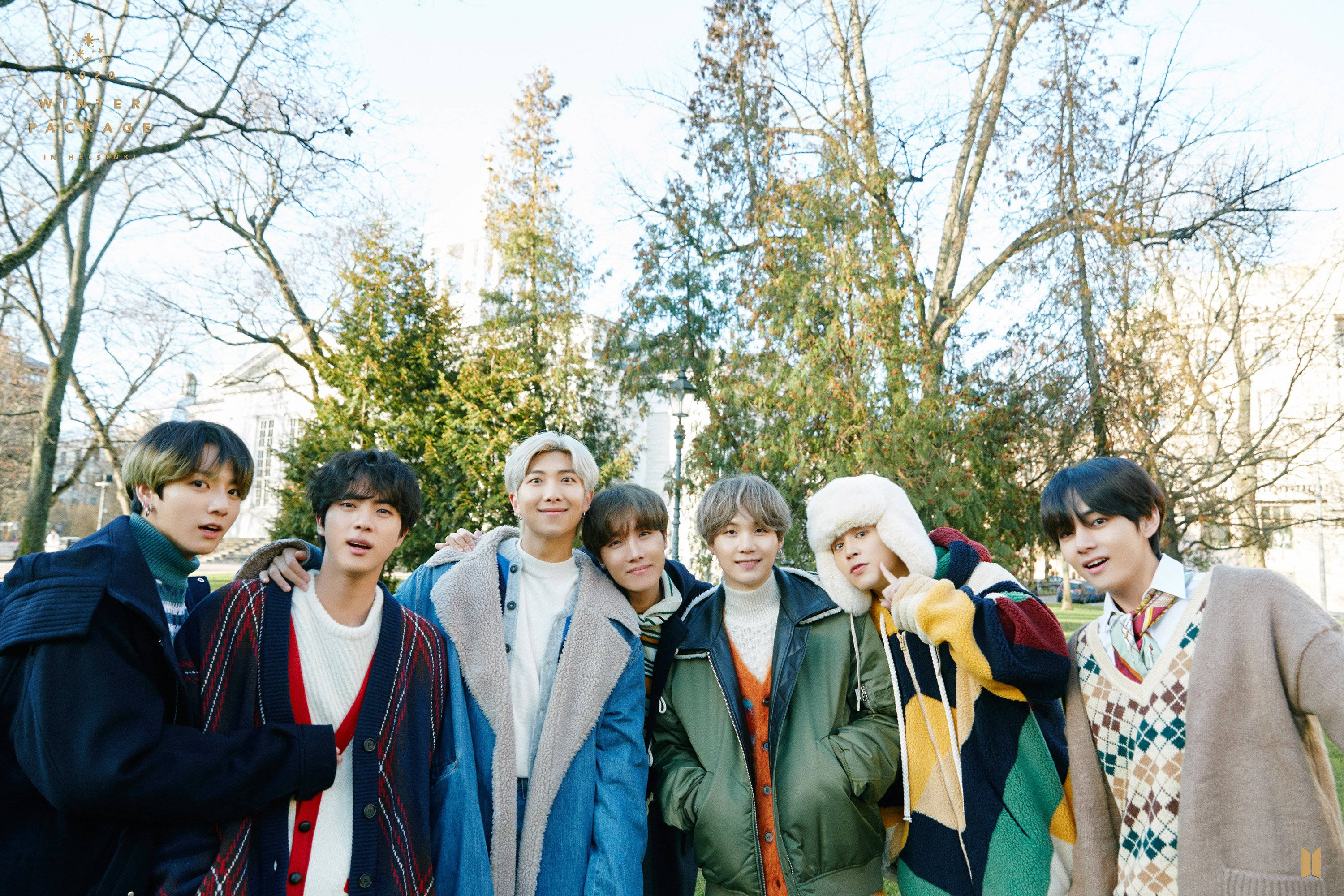 BTS 2021 桌布 壁紙 — 七個年輕男子穿著色彩繽紛的冬季外套，在陽光明媚的公園裡一起擺姿勢，背景是光禿禿的樹木和常青植物。