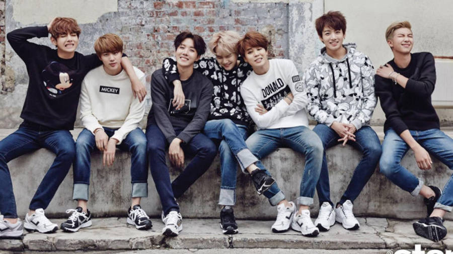 BTS 桌布 壁紙 — 七個年輕男子身穿休閒街頭服飾，一起坐在混凝土階梯上，背景是風化的磚牆，對著鏡頭溫暖微笑。