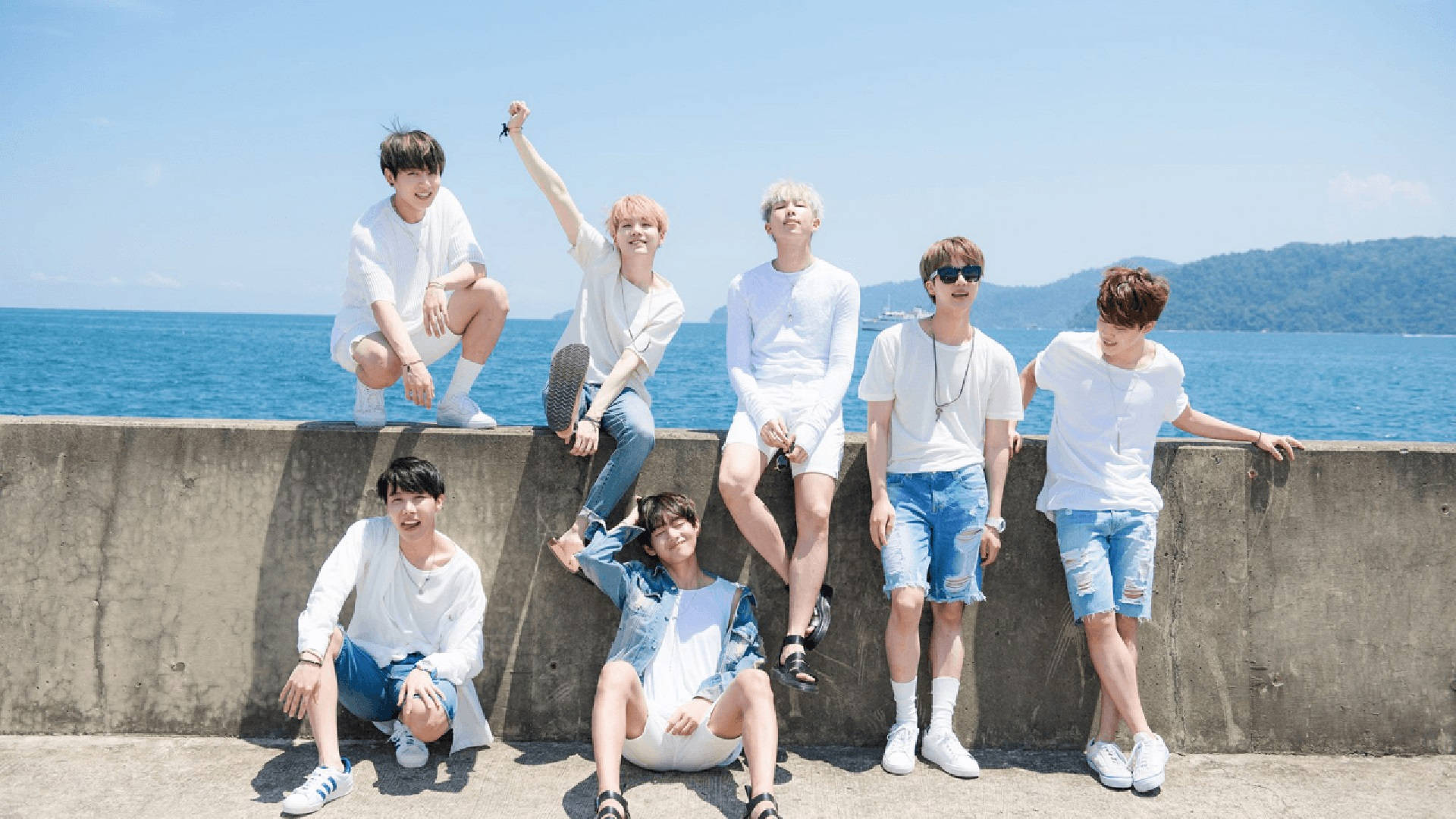 BTS 桌布 桌布 壁紙 — 七個年輕男子身穿白色上衣和牛仔短褲，在明亮的藍色海洋和天空背景下，在混凝土海堤上開心地擺姿勢。