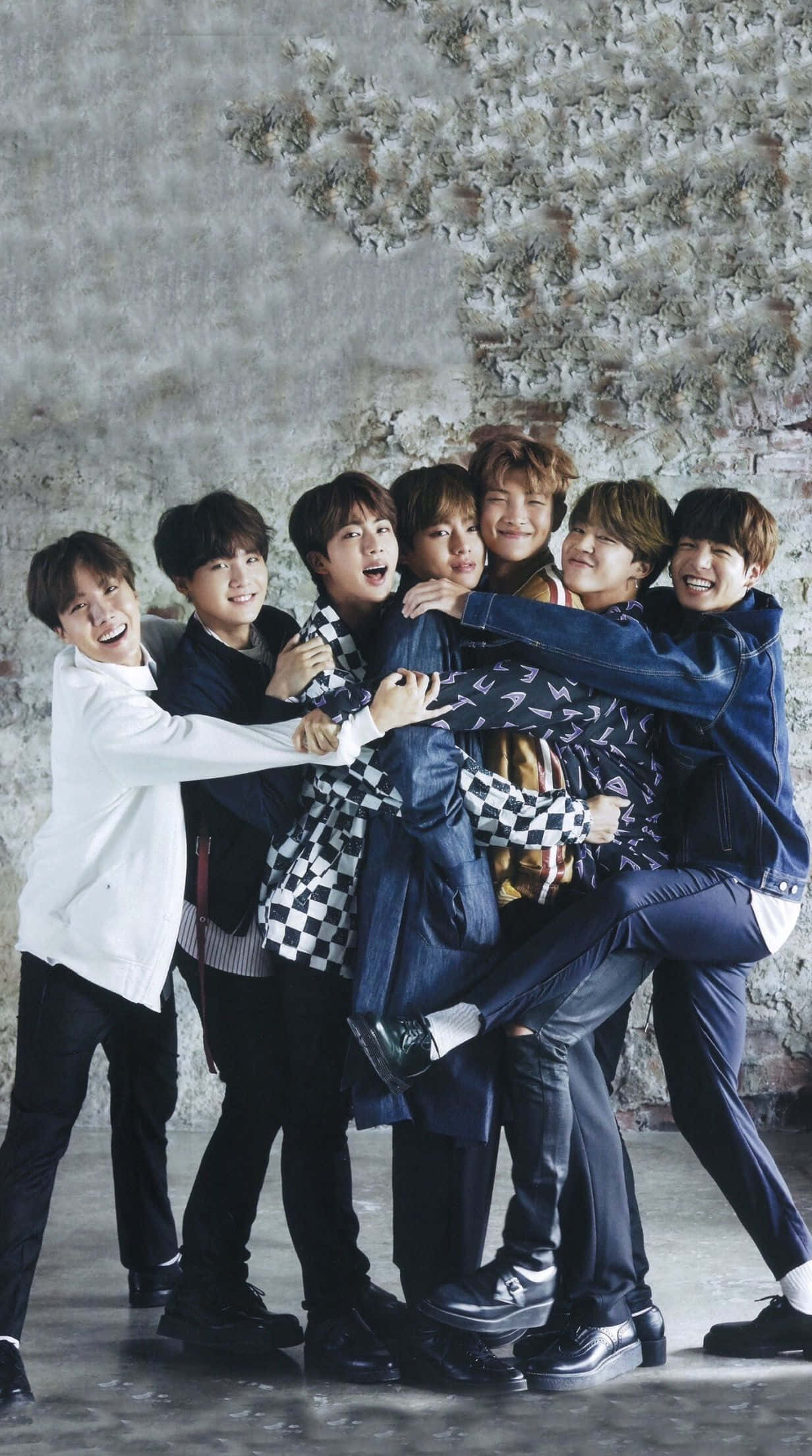 BTS 桌布 壁紙 — 七個微笑的年輕男子穿著休閒服裝，擠在一起靠著一面有紋理的灰色磚牆。