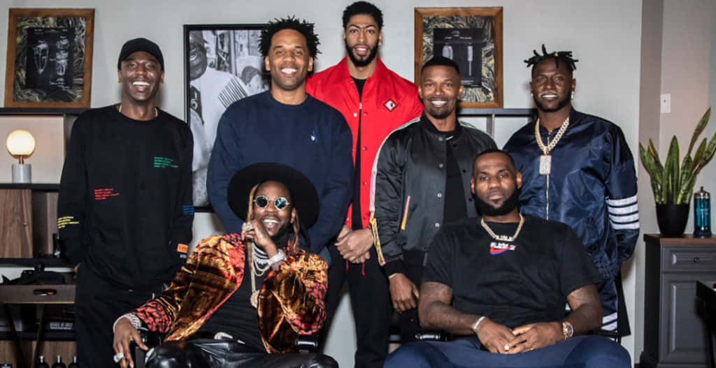 2 Chainz 桌布 壁紙 — 七個微笑的男人穿著時尚街頭服飾，在一個有裱框藝術品和植物的現代客廳裡一起擺姿勢。