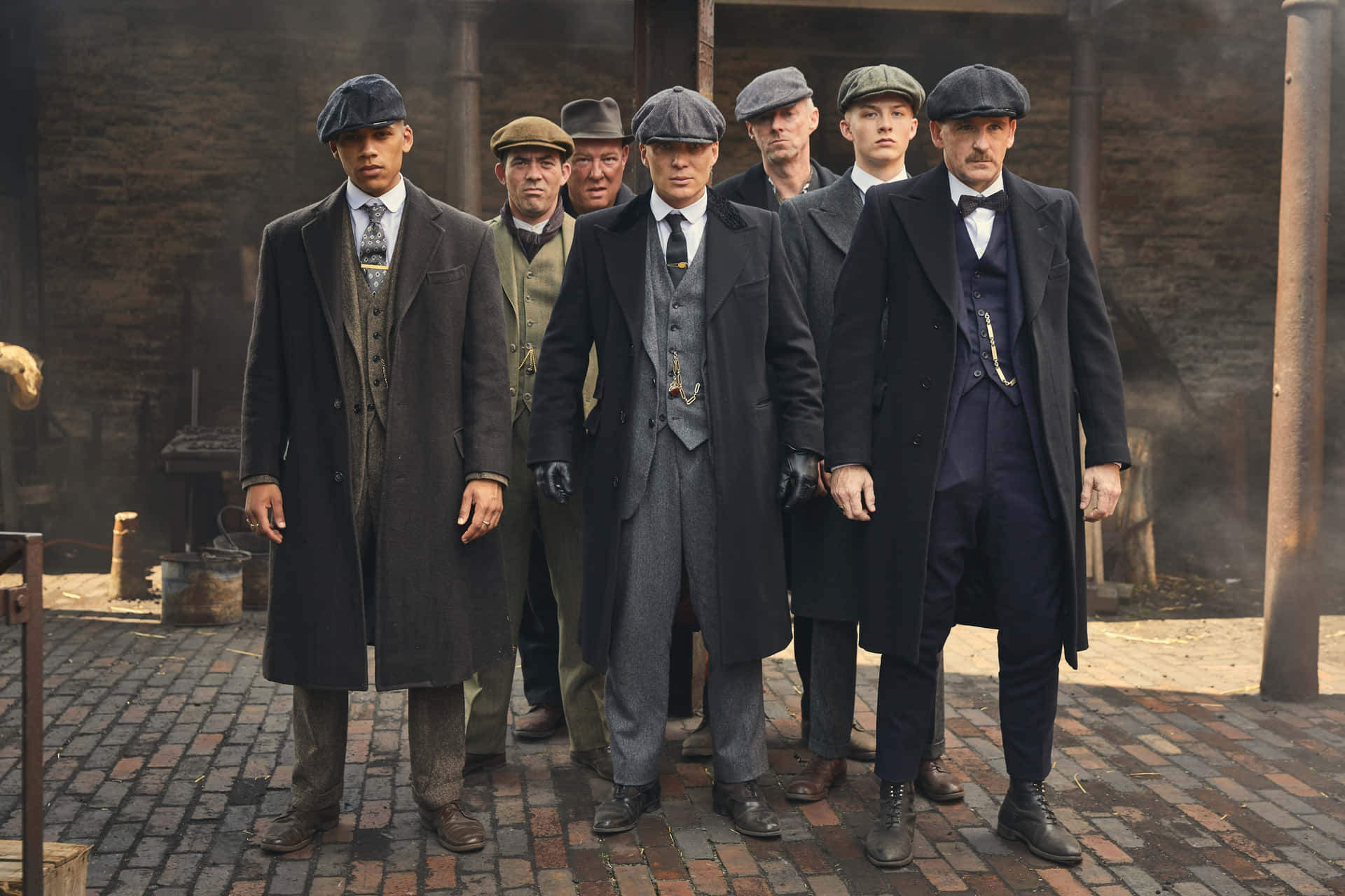 七個身穿復古西裝和帽子的男人，在有煙霧的磚砌庭院中站立。 — Peaky Blinders 壁紙