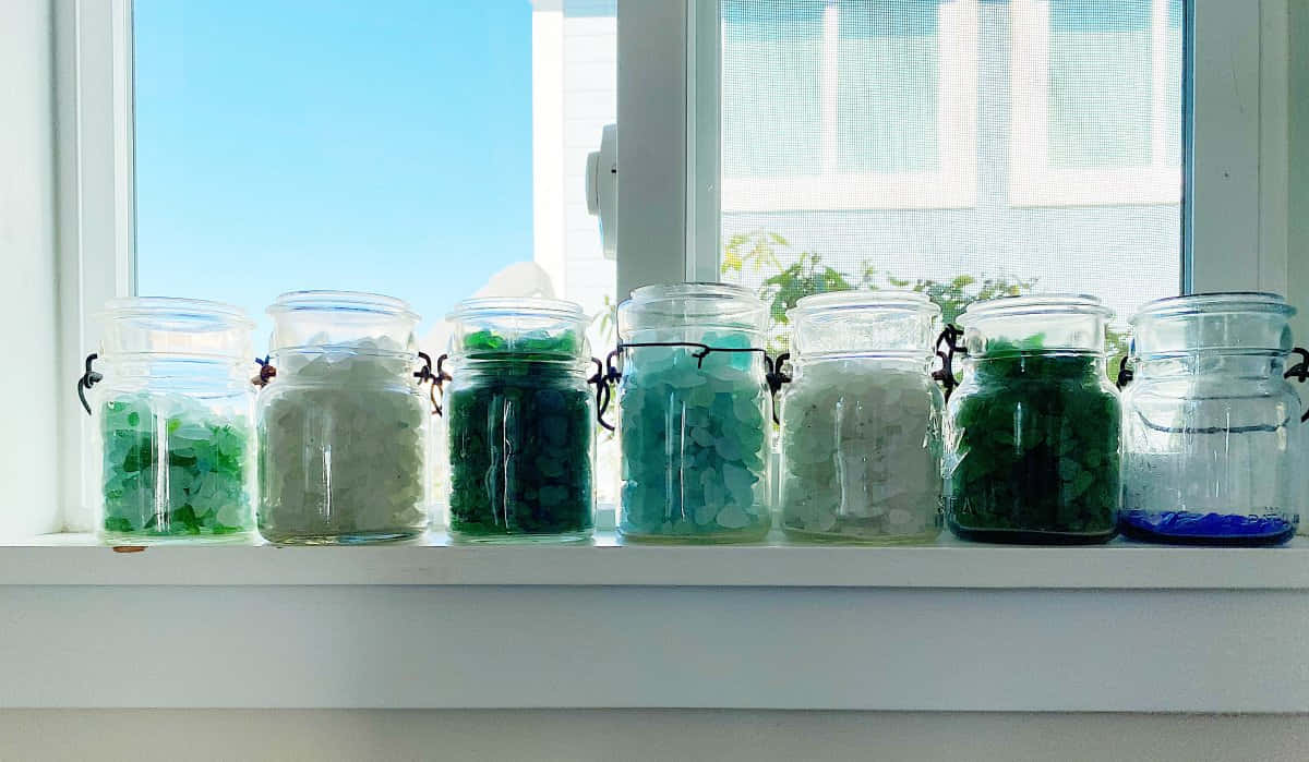 Sea Glass 桌布 壁紙 — 七個玻璃罐裝滿了各種綠色、藍色和白色的石頭，整齊地放在一個明亮的白色窗台上，沐浴在自然光中。