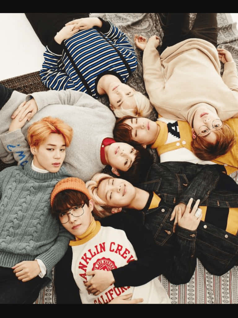 BTS 桌布 壁紙 — 七個穿著休閒服的年輕男子，躺在有圖案的地毯上圍成一個圓圈，放鬆地抬頭看著相機。