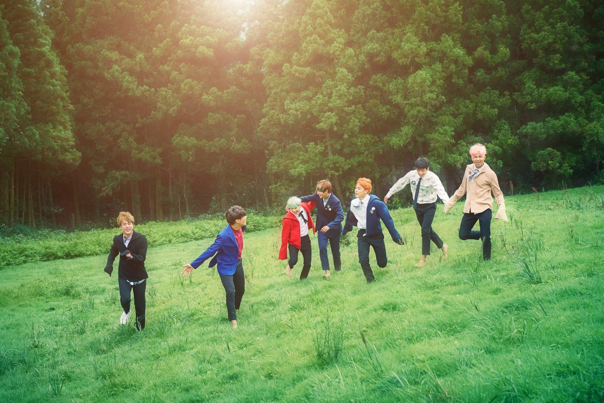 BTS 桌布 壁紙 — 七個穿著休閒服裝的人愉快地跑過一片陽光明媚的草地，周圍是茂密的綠樹。