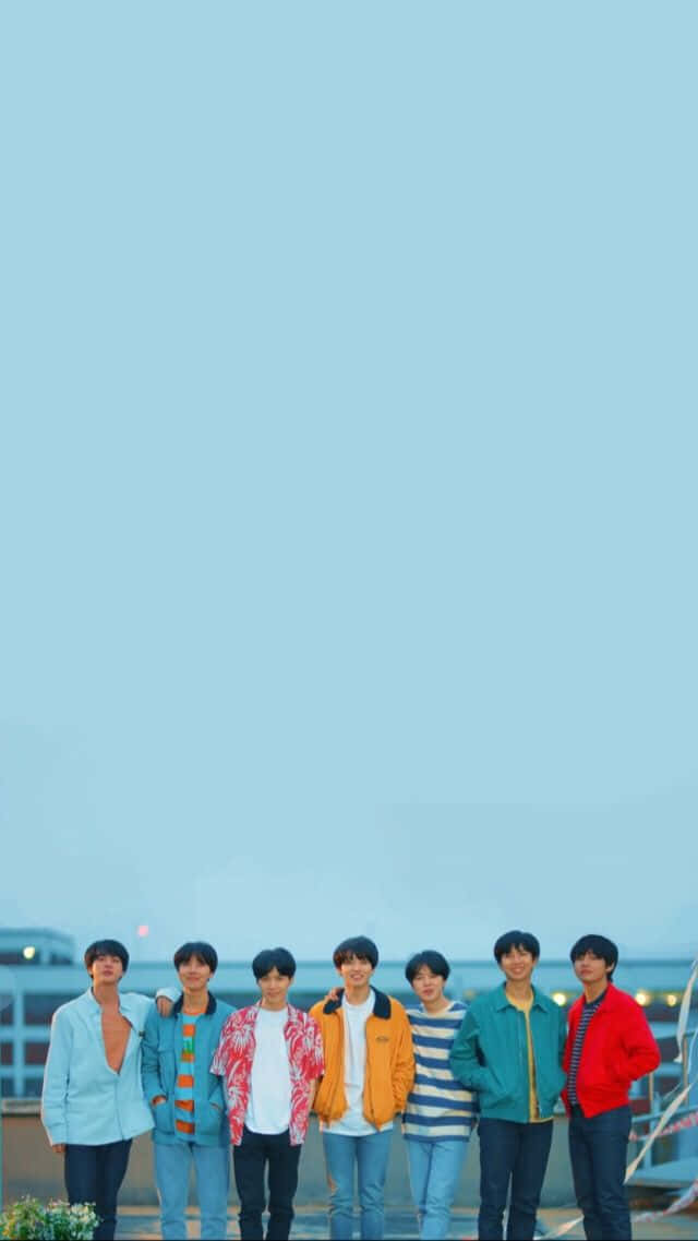 BTS 桌布 壁紙 — 七個穿著彩色外套的年輕男子在黃昏時分站在屋頂上，背景是廣闊的淡藍色天空。