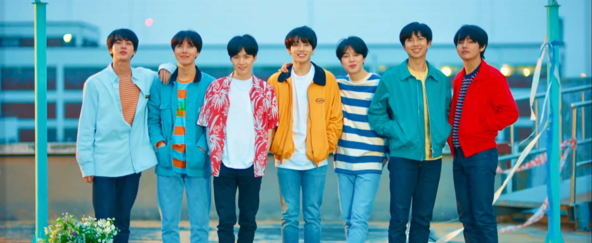 Bts Mac 桌布 壁紙 — 七個穿著彩色外套的年輕男子在黃昏時分一起站在屋頂上，背後是城市景觀。